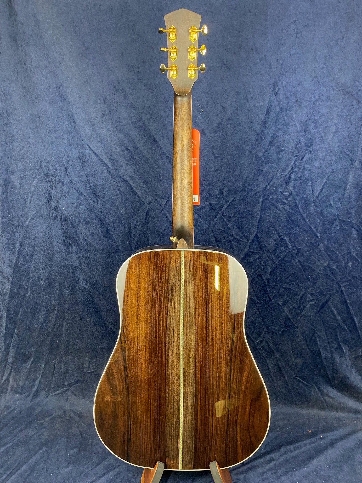 The back of an acoustic guitar with a glossy, dark wood finish and a lighter wood center stripe, gold tuners, and a red tag on the neck against a dark backdrop.
