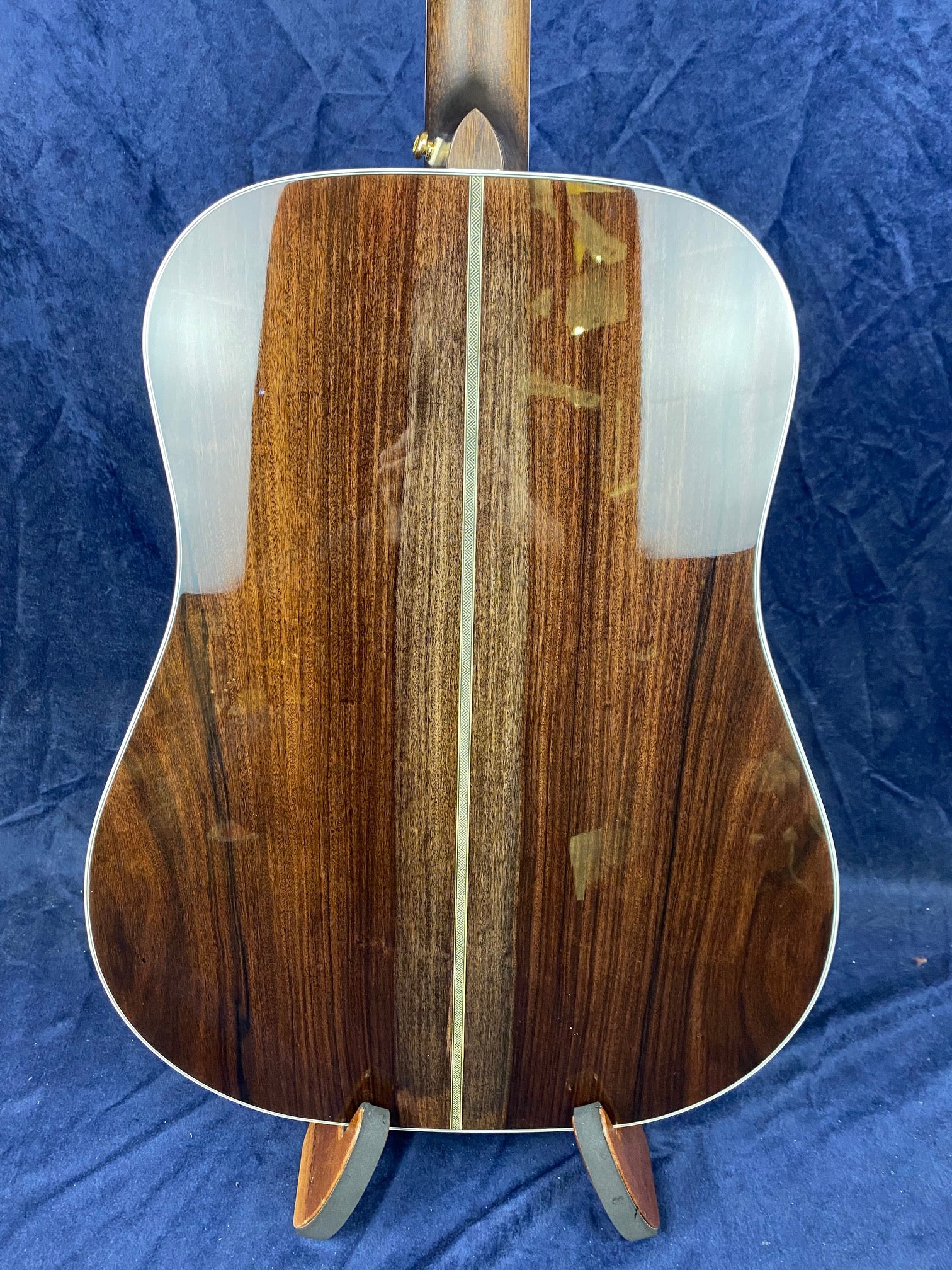 Acoustic guitar back with dark wood, a lighter wood center stripe, and patterned inlay; set on a stand against a blue textured background.