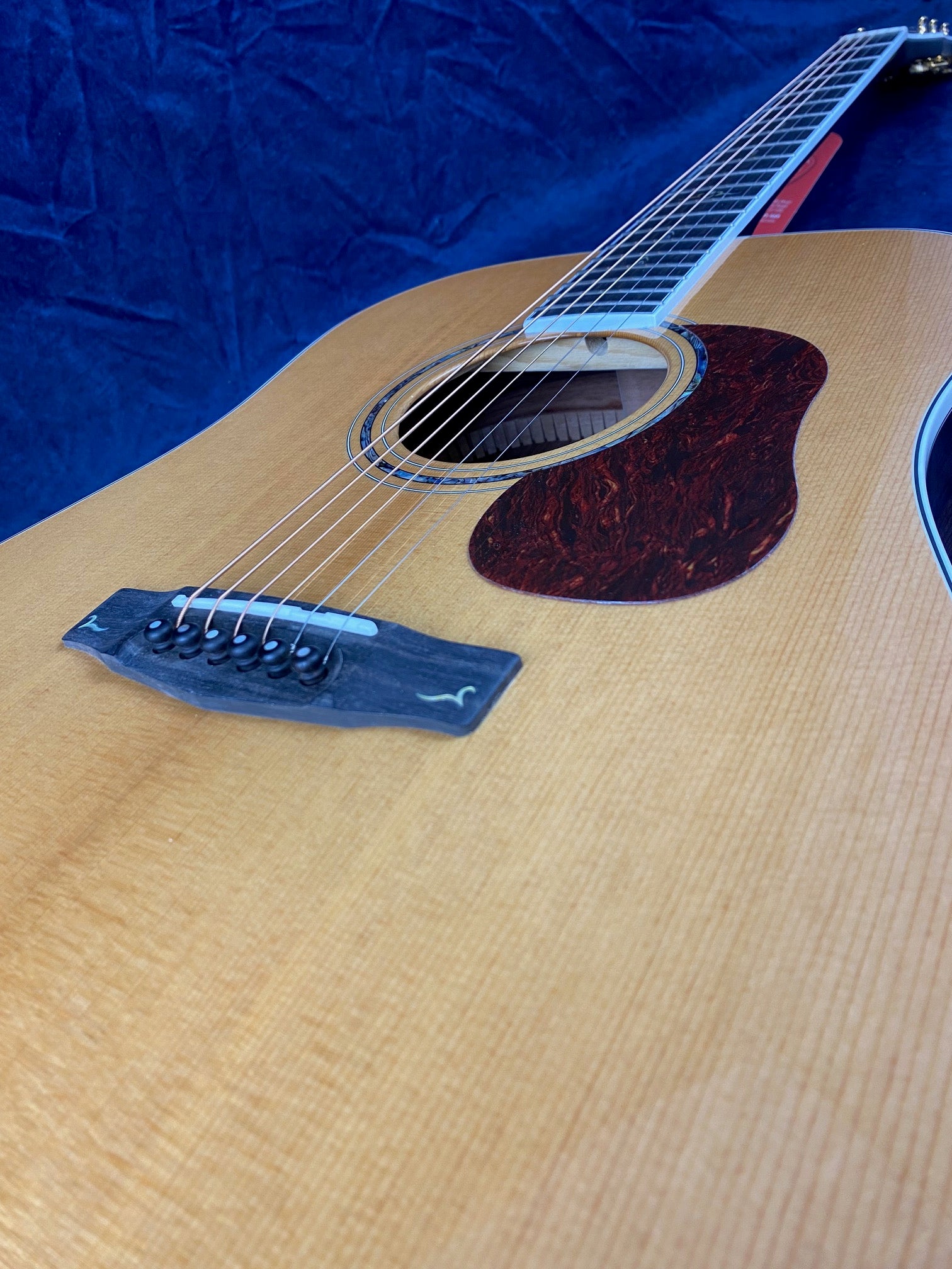 Light wood acoustic guitar with a dark tortoiseshell-style pickguard, six strings, and a black bridge, shown lying on a blue background.