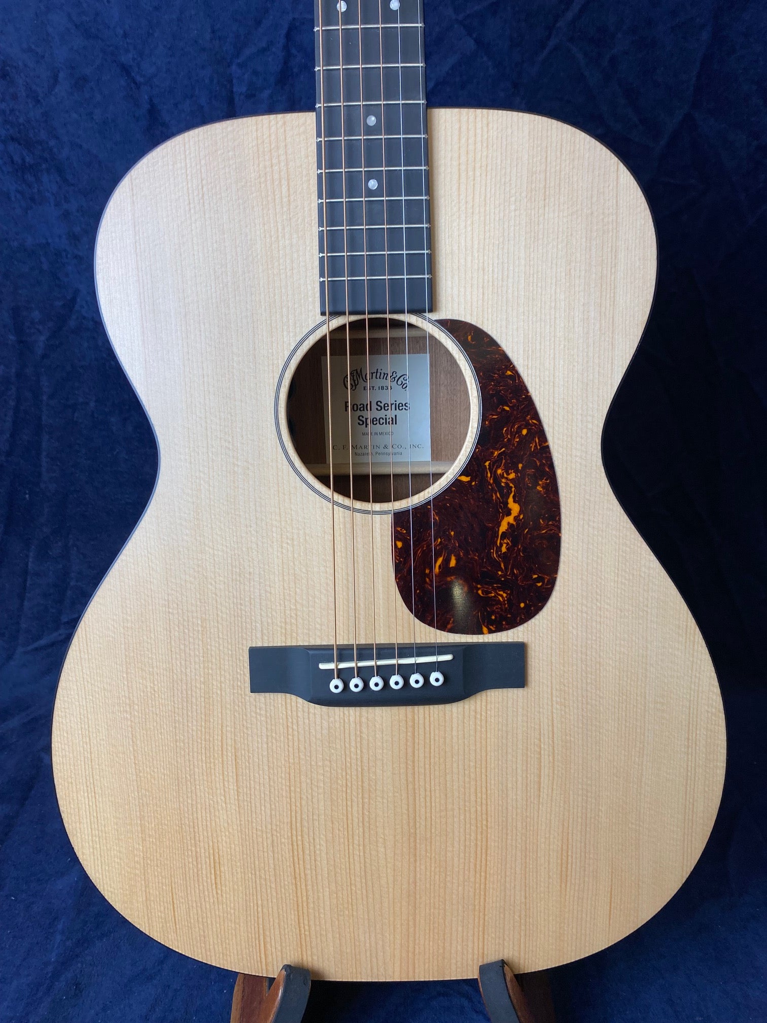 Light wood acoustic guitar with a dark tortoiseshell-pattern pickguard, six strings, and a black bridge, displayed vertically against a dark background.