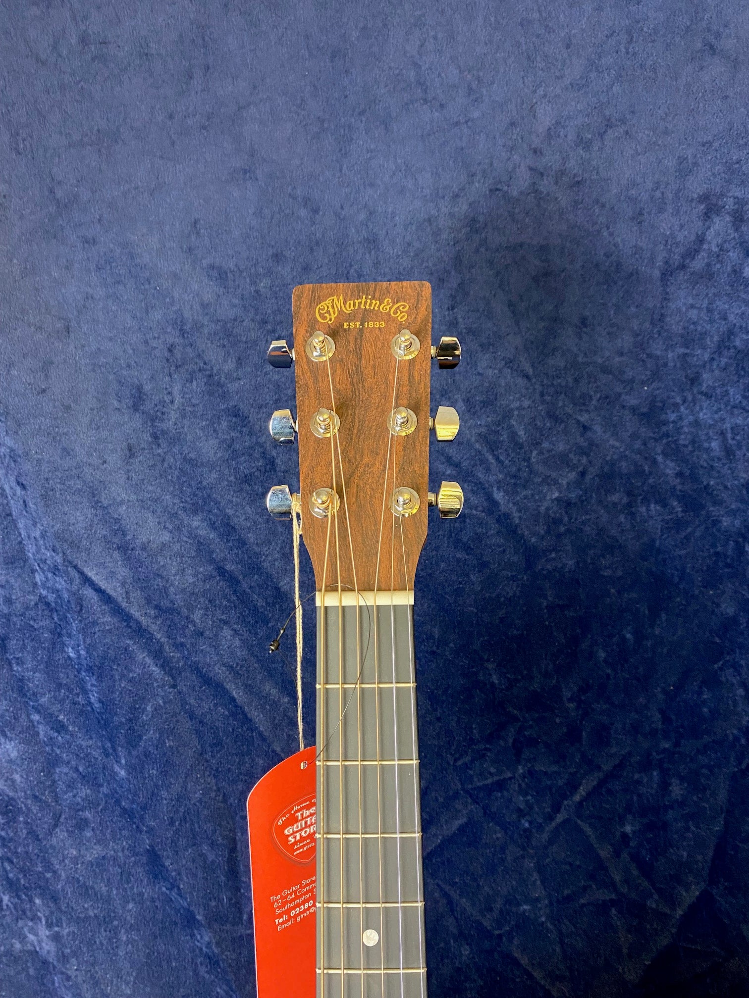 Martin & Co. acoustic guitar headstock with six tuning pegs, a loose string, and a red tag hanging from one tuner against a dark blue background.