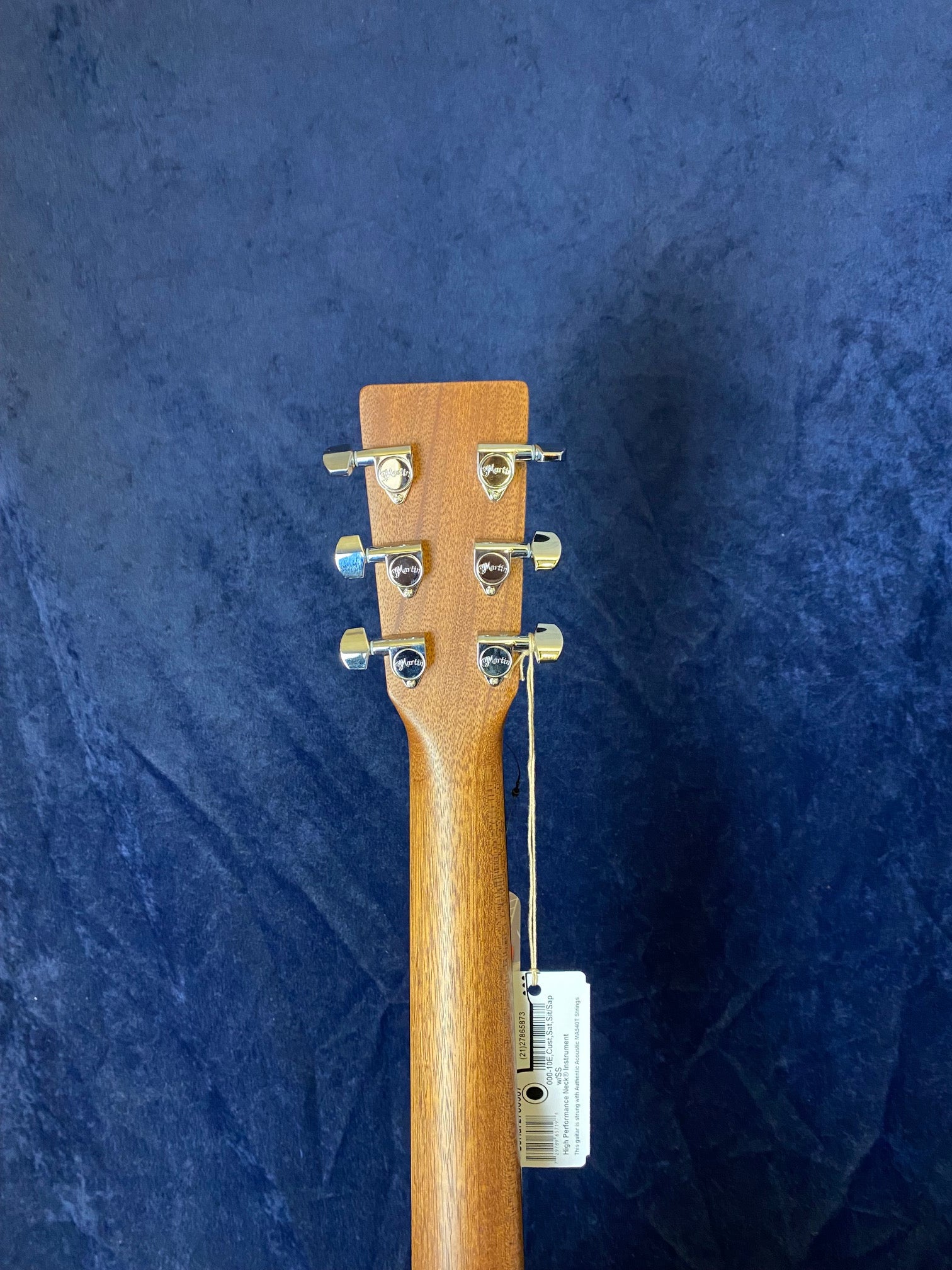 Wooden acoustic guitar neck and headstock shown from behind, with six silver tuning pegs and a price tag attached by a string.