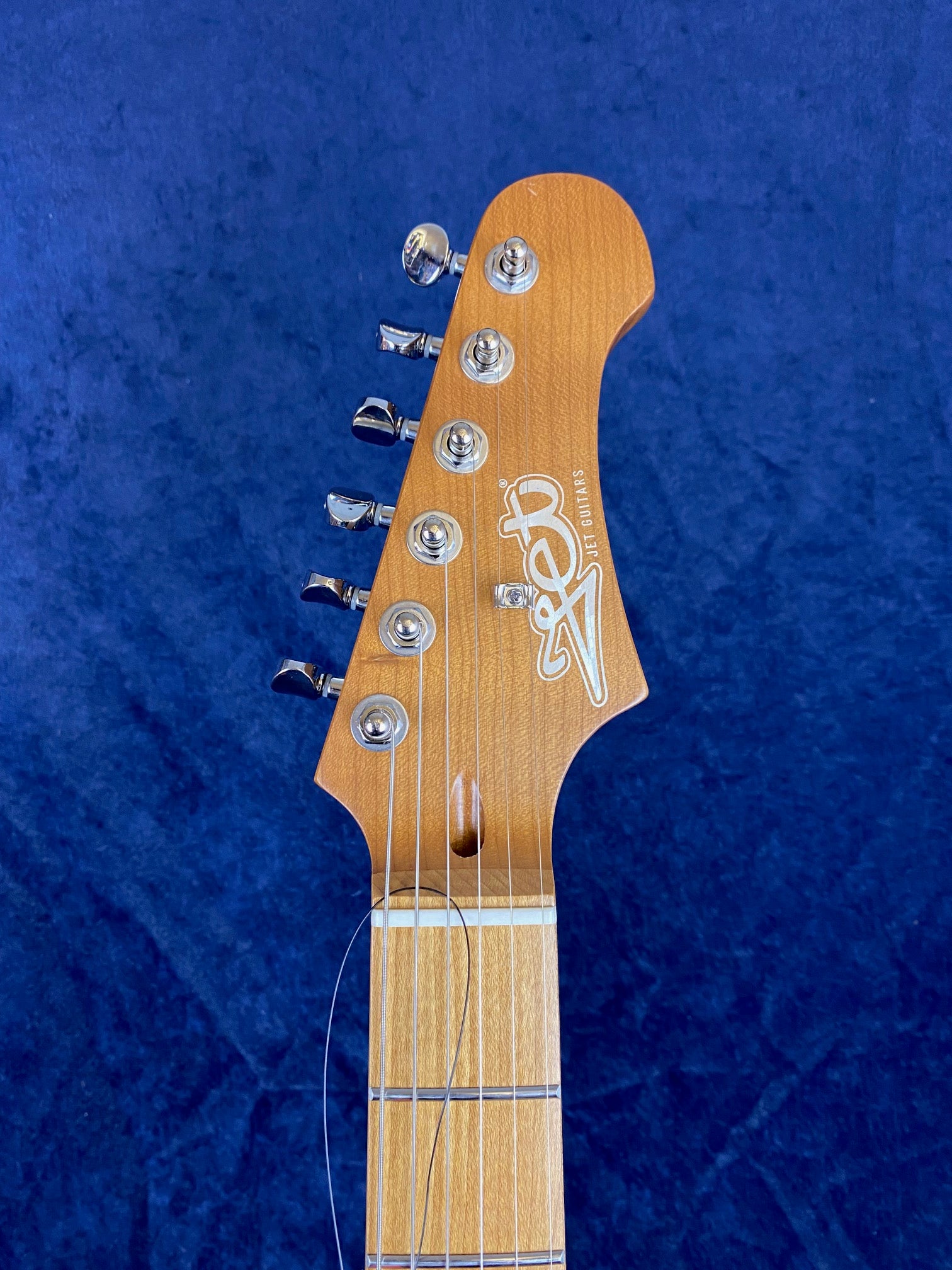 Light wood electric guitar headstock with six metal tuning pegs, "JET Guitars" logo in white, and unfinished string against a dark blue background.