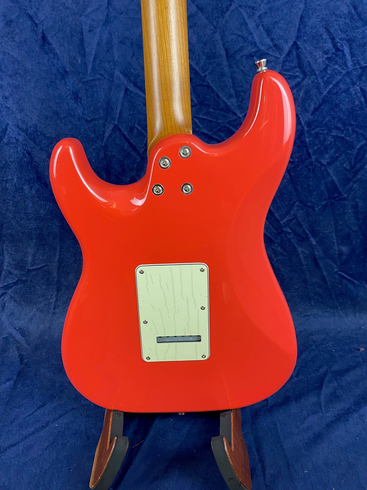The back of a bright red electric guitar with a natural wood neck and a cream-colored rectangular cover plate, shown against a dark blue fabric background.