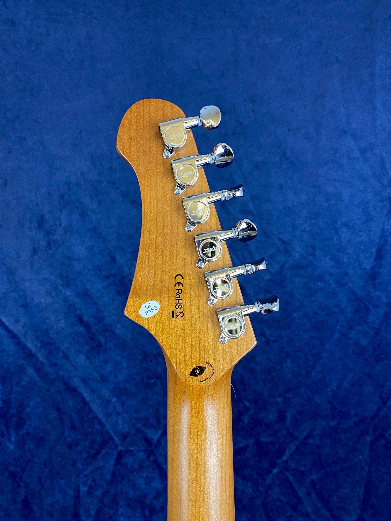 Six chrome tuning pegs on the back of a light wood electric guitar headstock, with quality control and certification stickers visible.