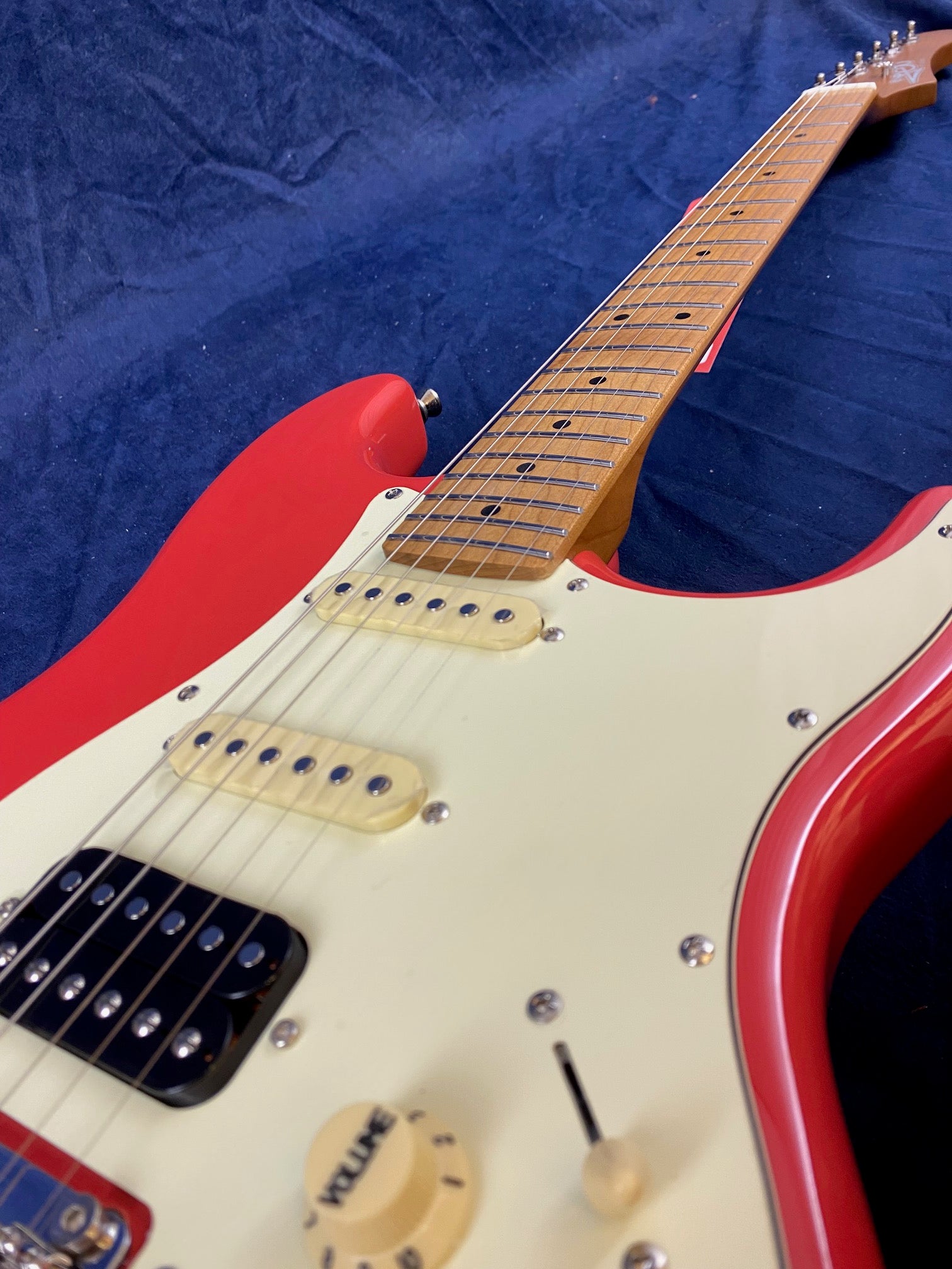 Red electric guitar with a white pickguard, three pickups, a maple neck, and a knob labeled "volume" on a dark blue fabric background.