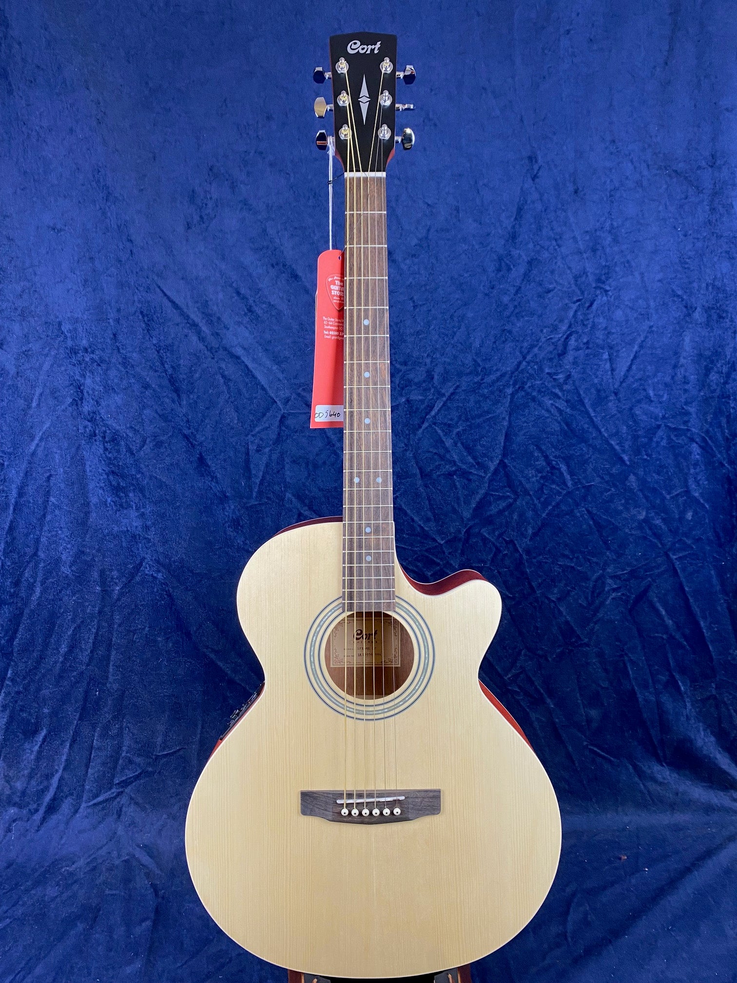 Cort acoustic guitar with a natural wood finish, cutaway body, six strings, and a red tag hanging from the tuning pegs against a blue background.