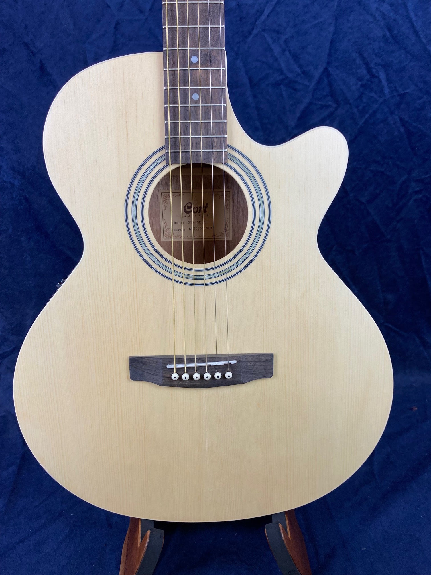 Light wood acoustic guitar with a cutaway body, six strings, and a dark wooden bridge, displayed upright against a dark blue background.