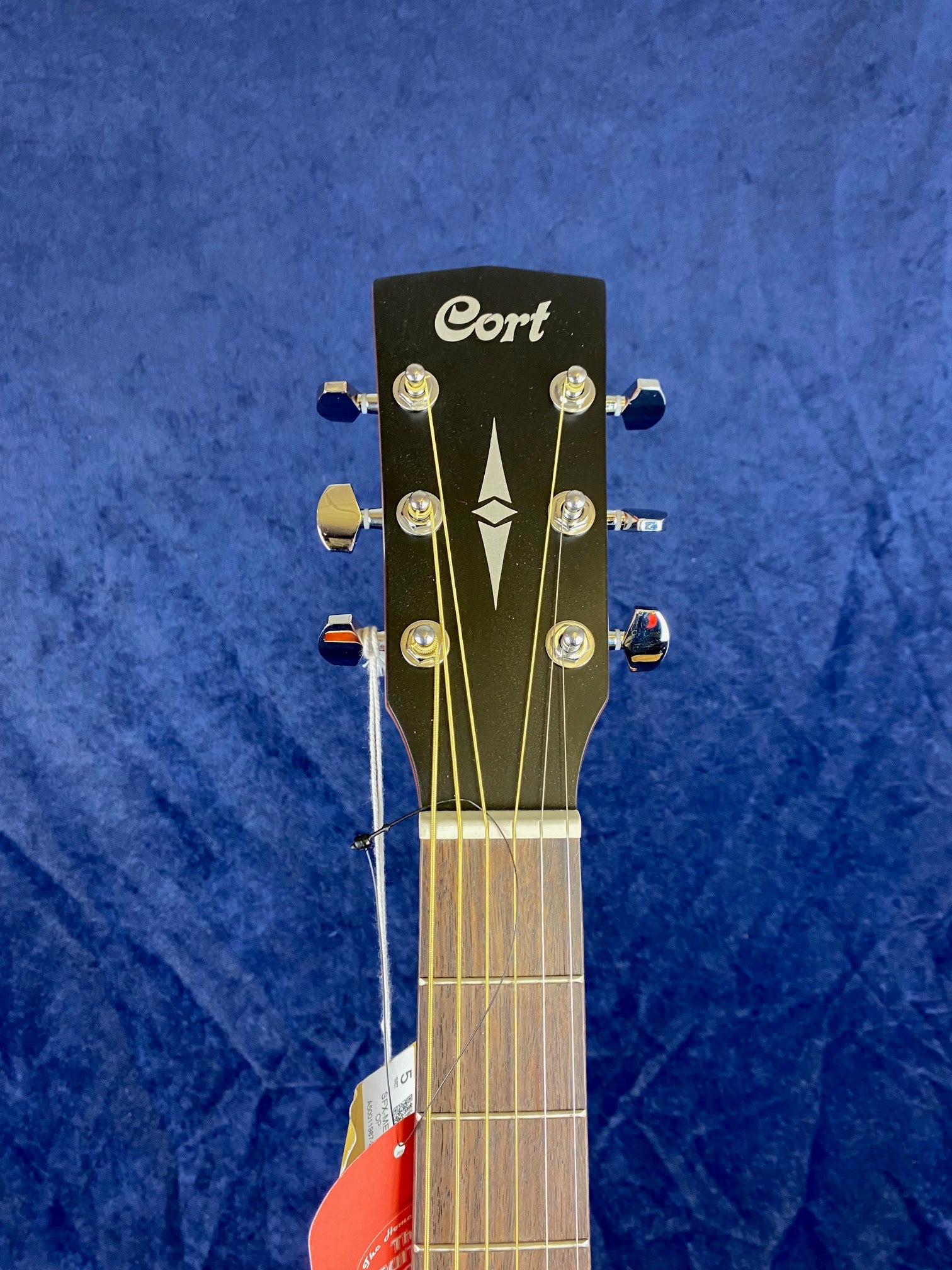 Cort acoustic guitar headstock with six tuning pegs, gold strings, and a white geometric inlay on a dark background.