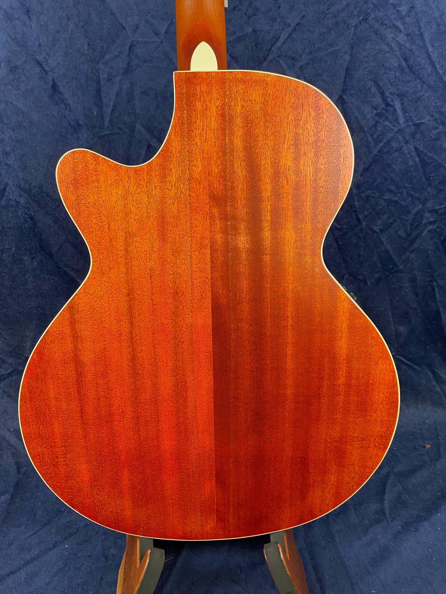 Back view of an acoustic guitar with a cutaway, rich reddish brown wood grain, and light binding along the edges, shown upright on a stand.