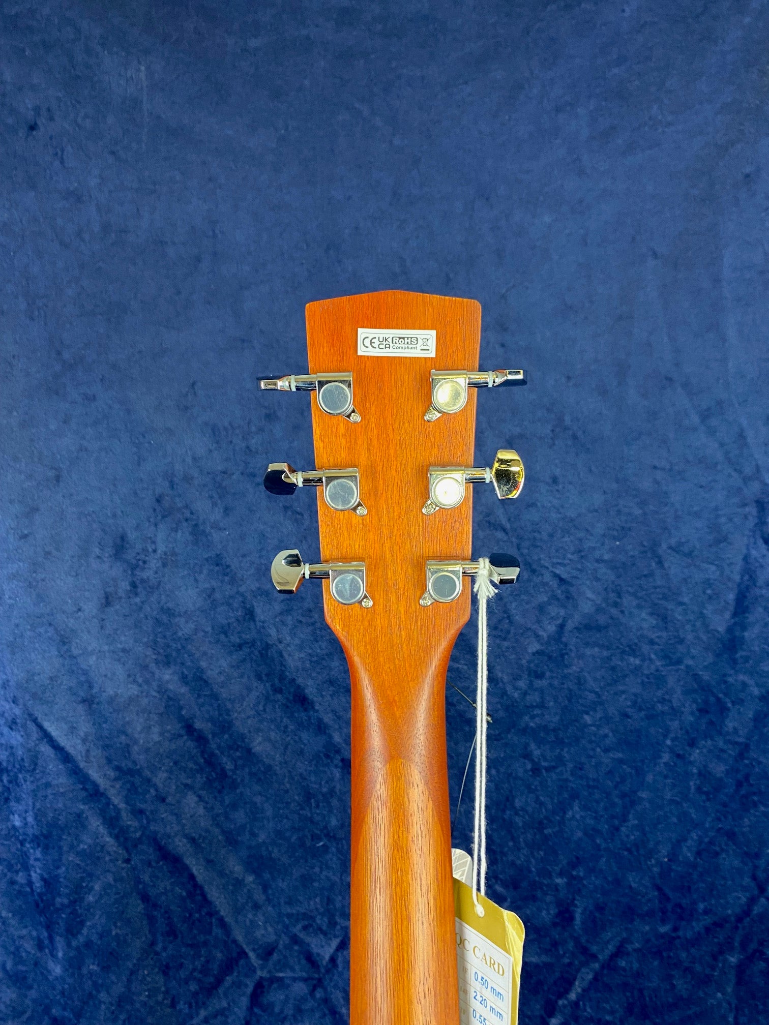 Back of an acoustic guitar headstock showing six chrome tuning pegs, a certification sticker, and a tag hanging from the neck against a blue background.