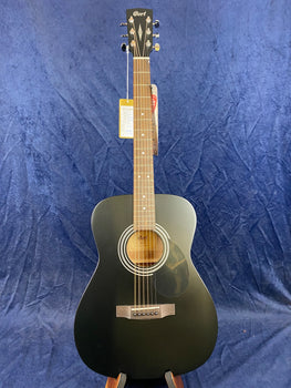 Matte black Cort acoustic guitar with six strings, light wood fretboard, and circular sound hole, displayed upright against a dark blue fabric background.
