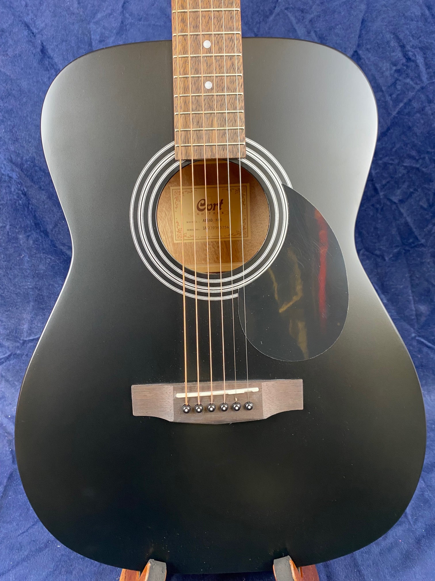 Matte black acoustic guitar with a round sound hole, white circular detailing, dark pickguard, and wooden bridge, displayed against a blue background.