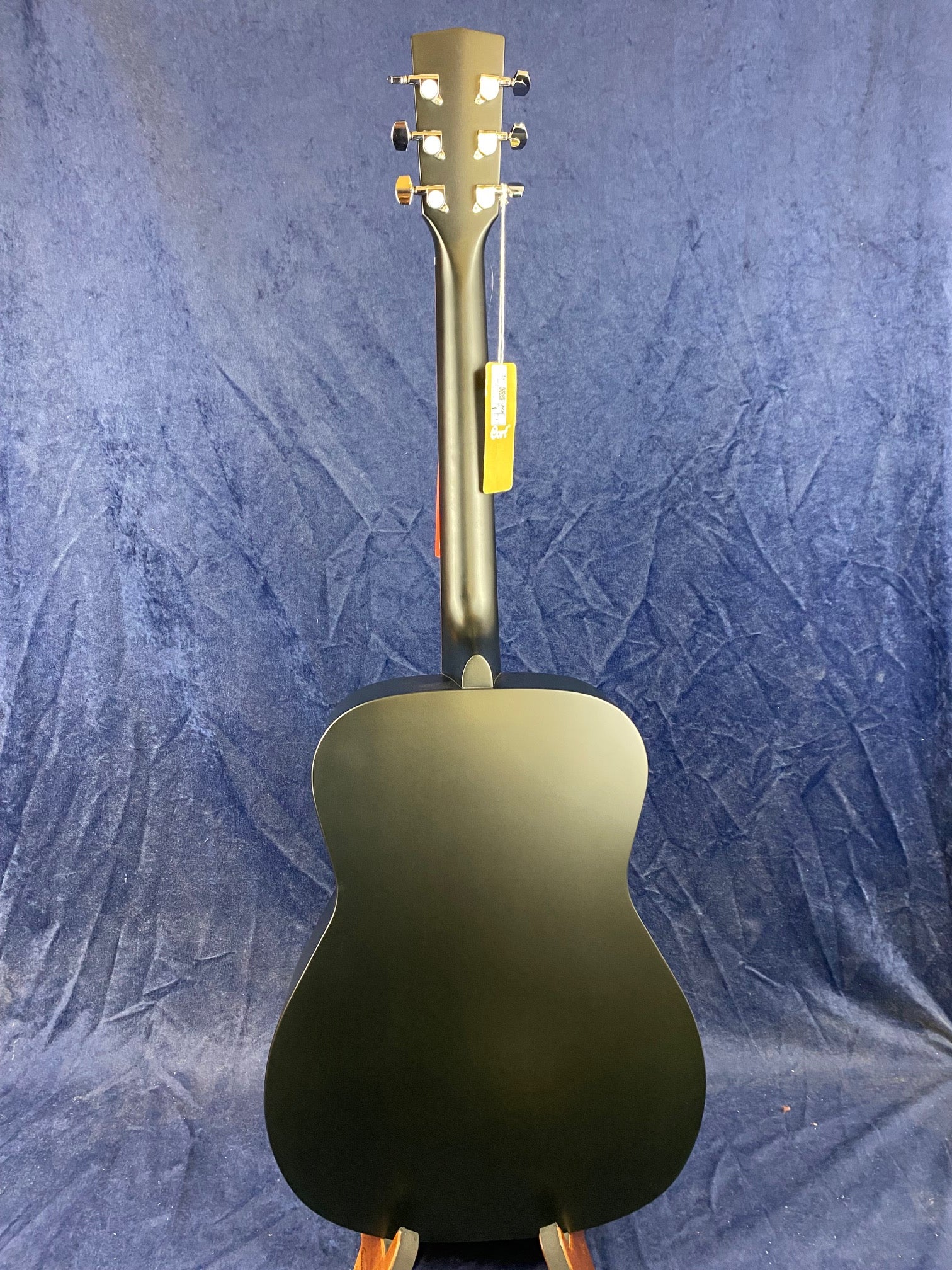 Black acoustic guitar shown from the back, with chrome tuning pegs and a tag attached to the headstock, set against a dark blue textured background.