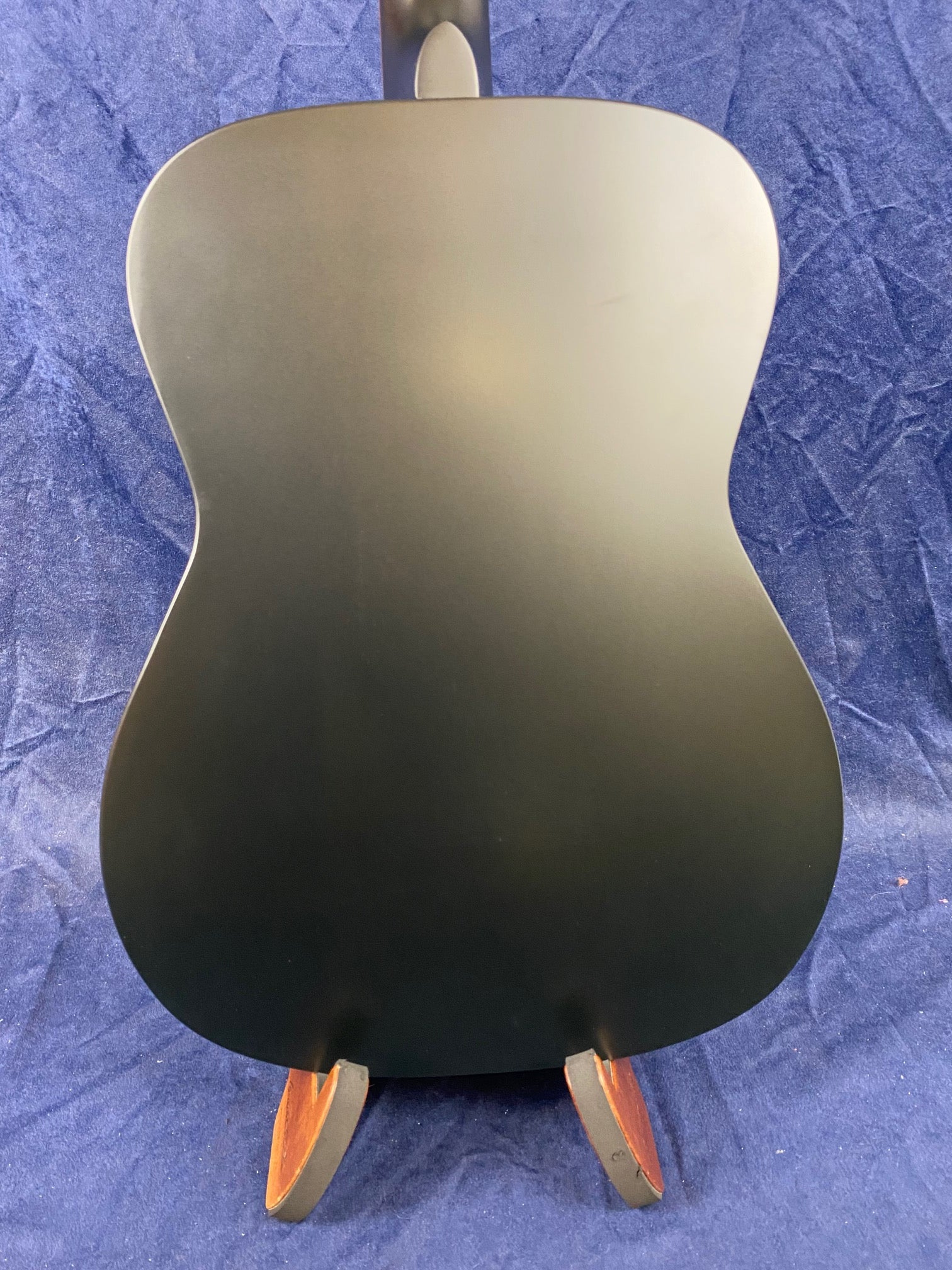 Matte black acoustic guitar back with smooth, unadorned finish, resting on a wooden stand against a textured blue fabric background.