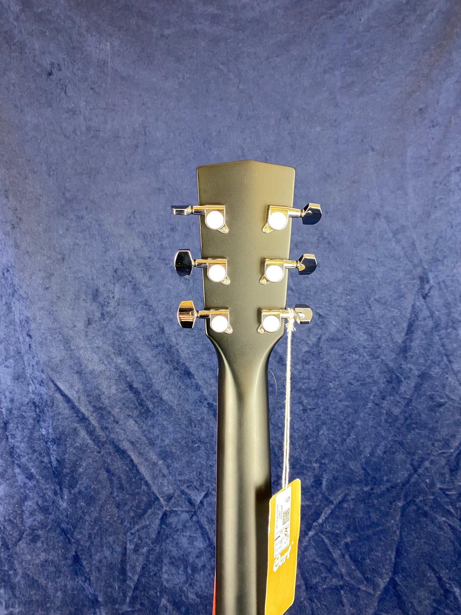 Rear view of an acoustic guitar headstock with six silver tuning pegs and a hanging cardboard tag against a dark blue fabric background.