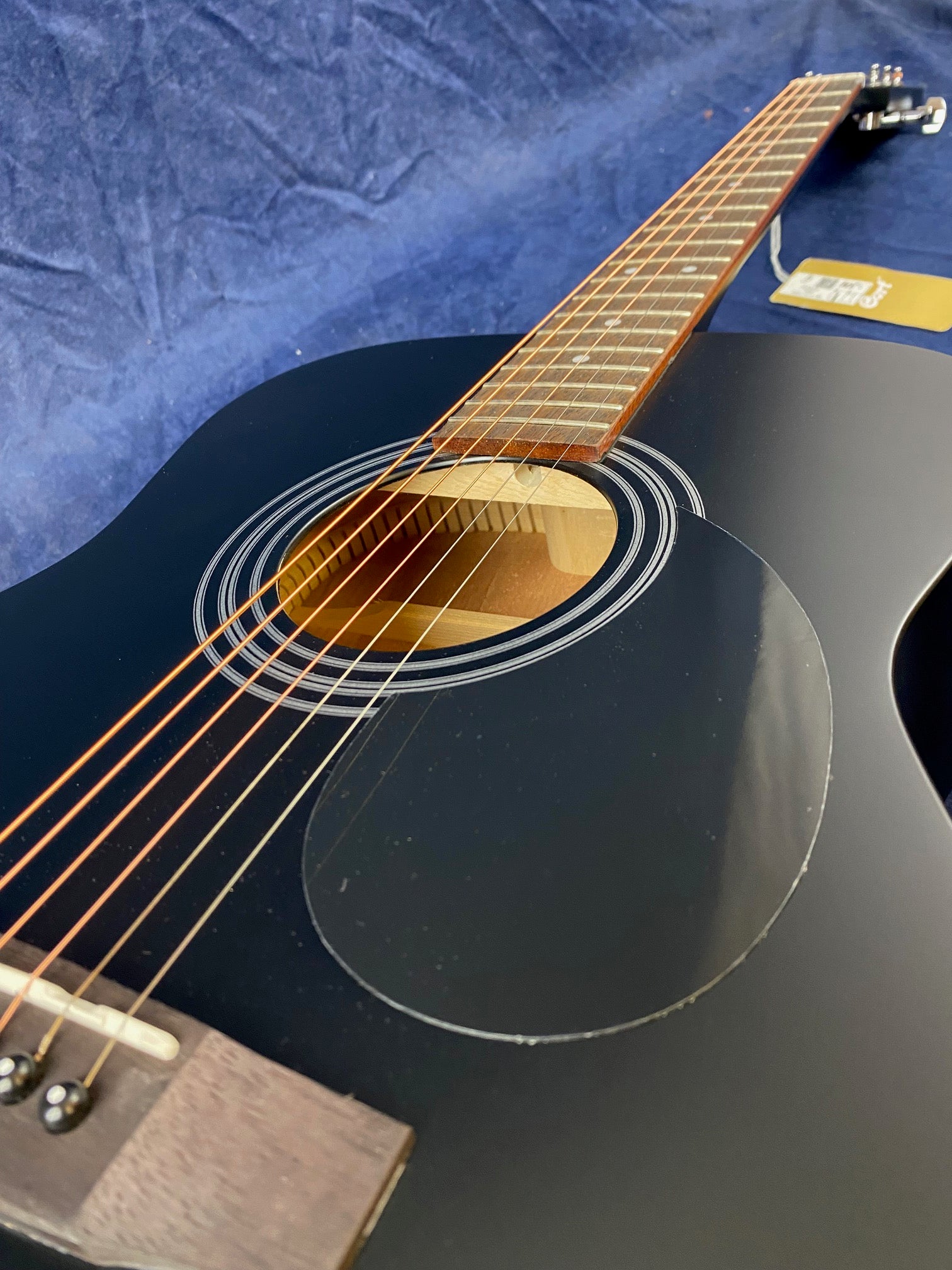 Black acoustic guitar with bronze strings, wooden fretboard, and a circular sound hole, viewed close up along the neck against a blue background.