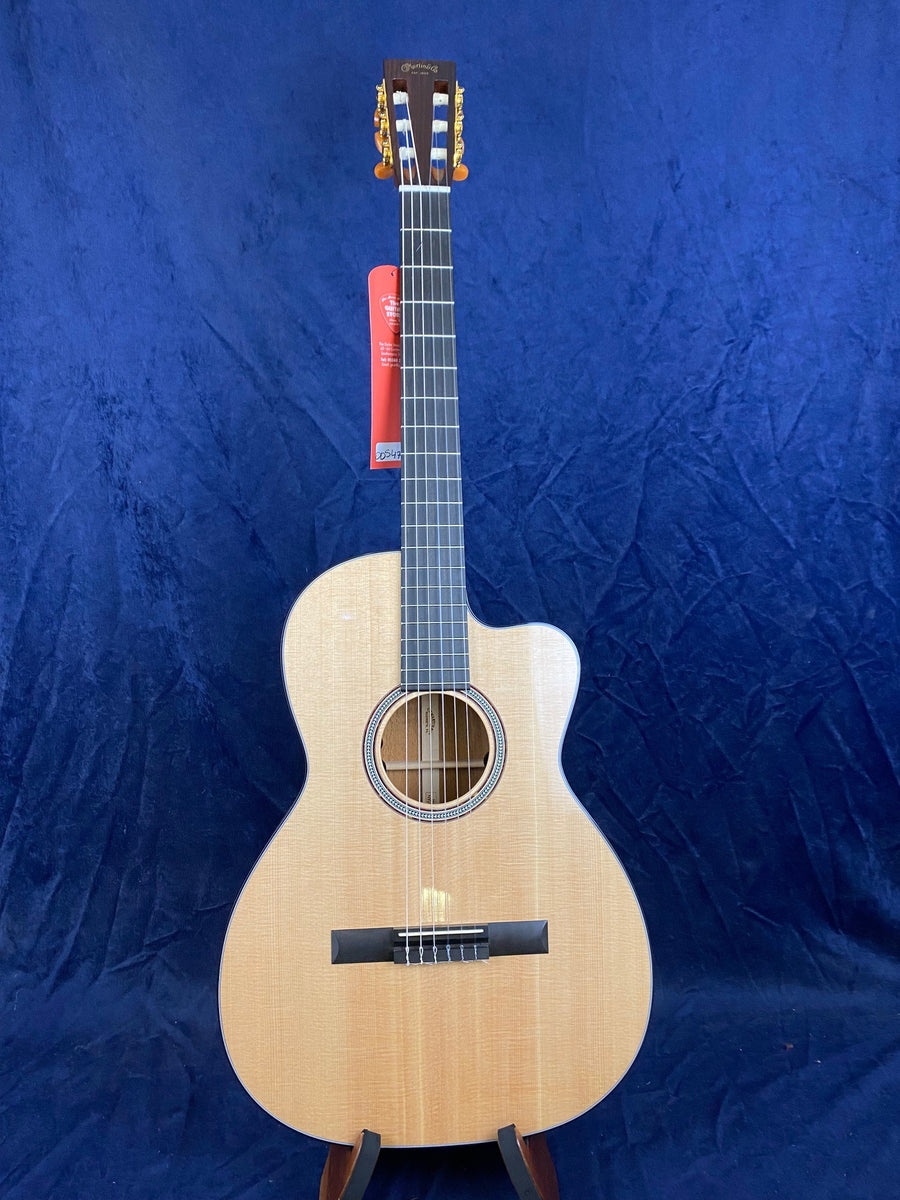 Natural-finish acoustic guitar with nylon strings, a cutaway body for easier high-fret access, and visible tuning pegs, set against a blue background.