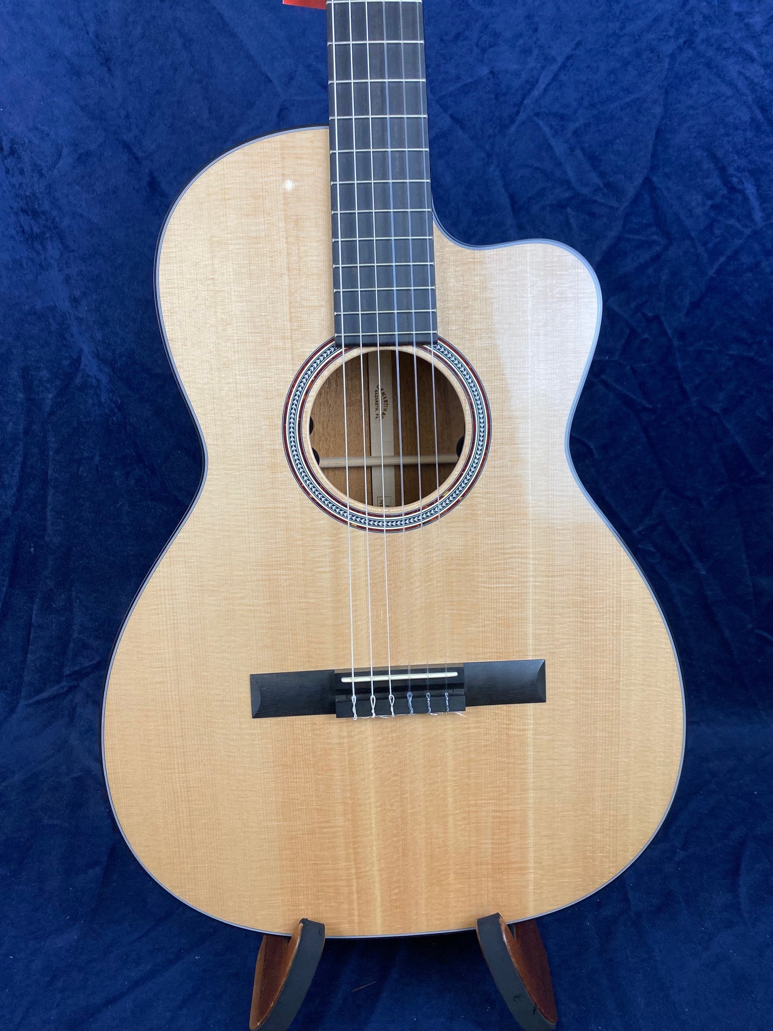 Light wood acoustic guitar with a cutaway body, black fretboard, six strings, and decorative rosette around the soundhole, displayed on a stand.