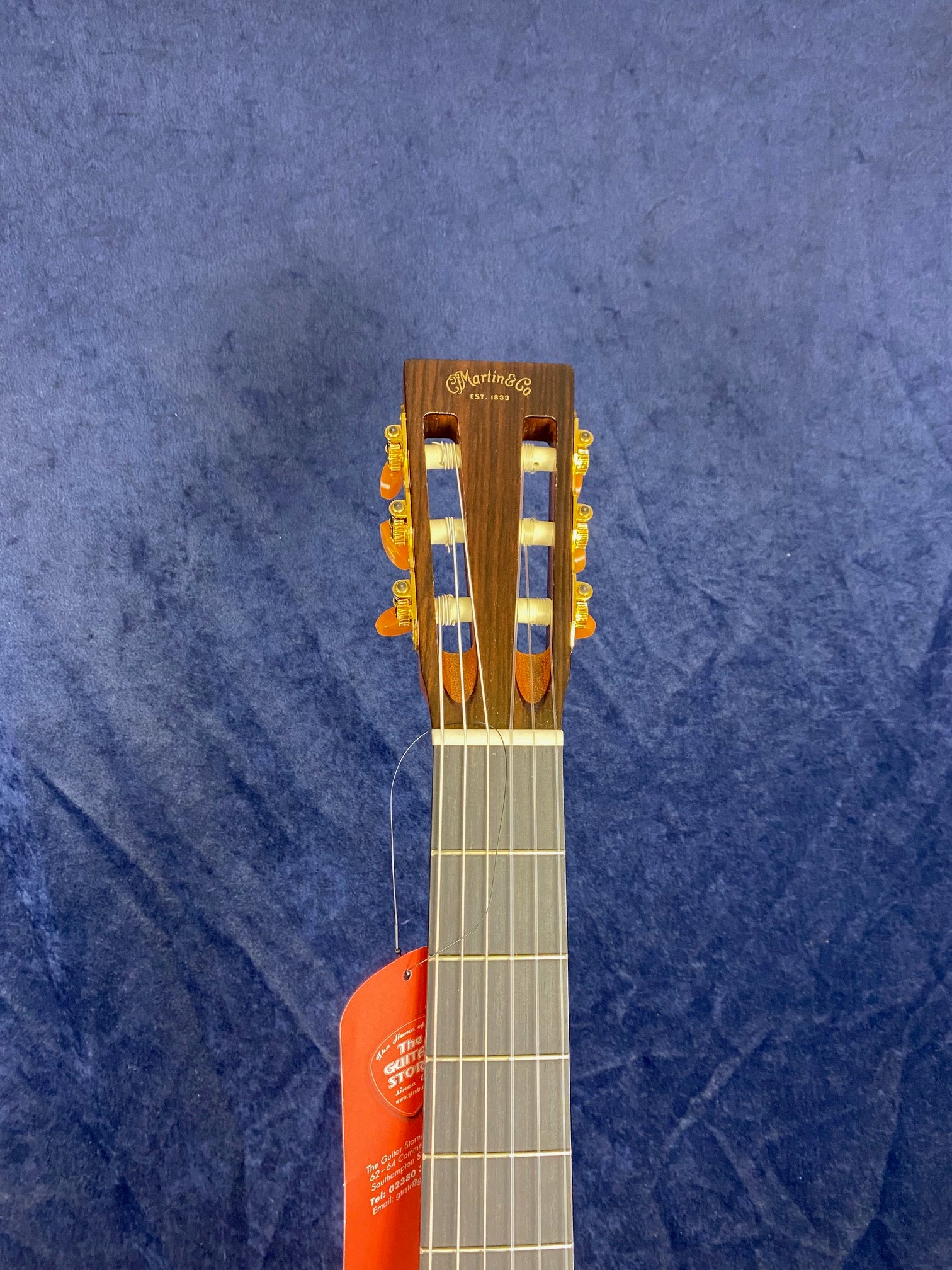 Headstock of a Martin & Co acoustic guitar with gold tuners and a red tag hanging from the neck, set against a dark blue background.