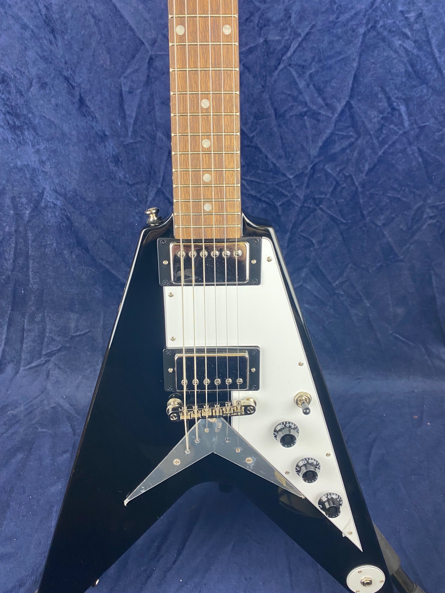 V-shaped black electric guitar with a white pickguard, six strings, two humbucker pickups, three control knobs, and a switch on a blue background.