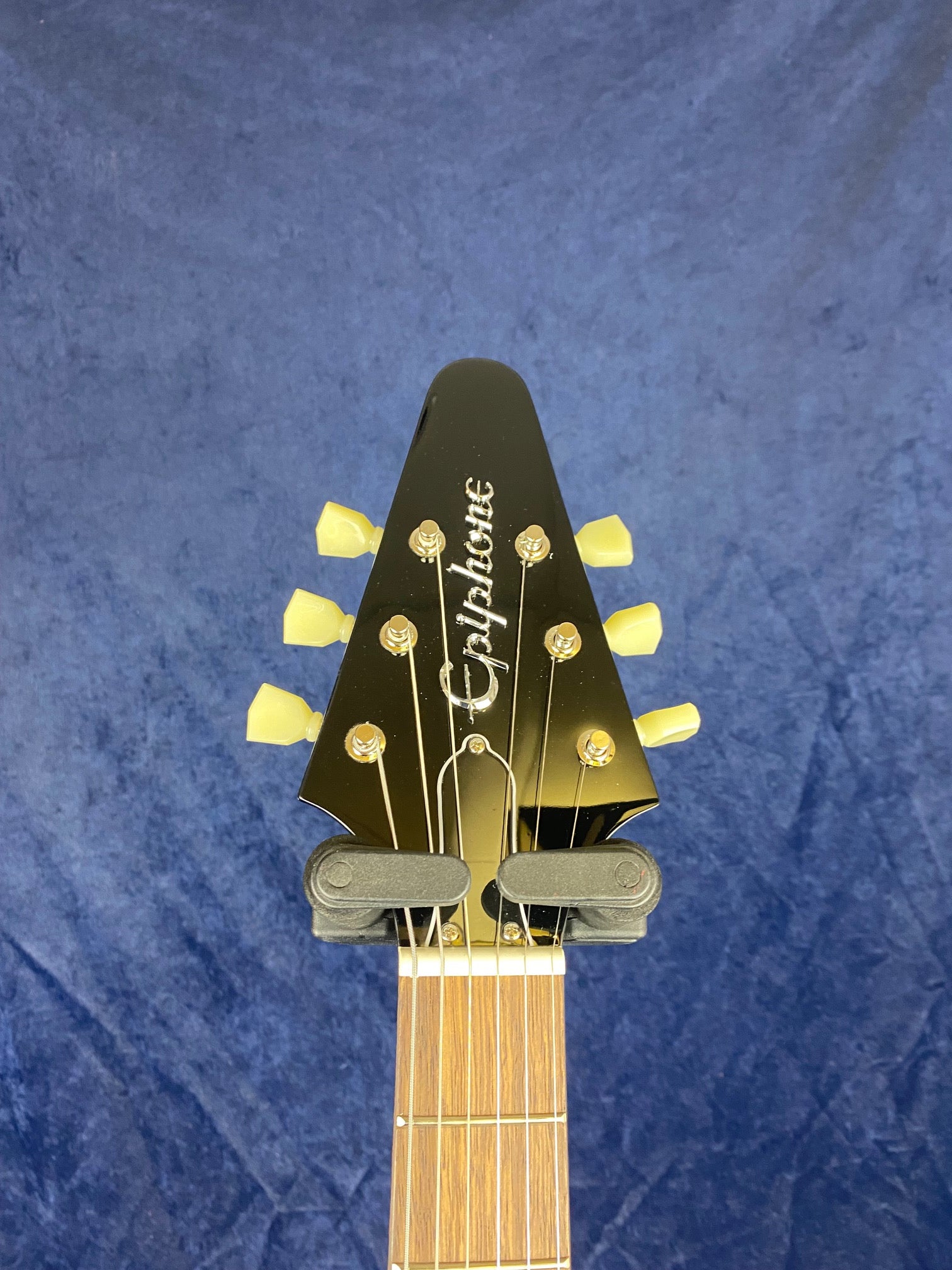 Black Epiphone electric guitar headstock with six cream tuners, silver logo, and a brown fretboard on a blue background.