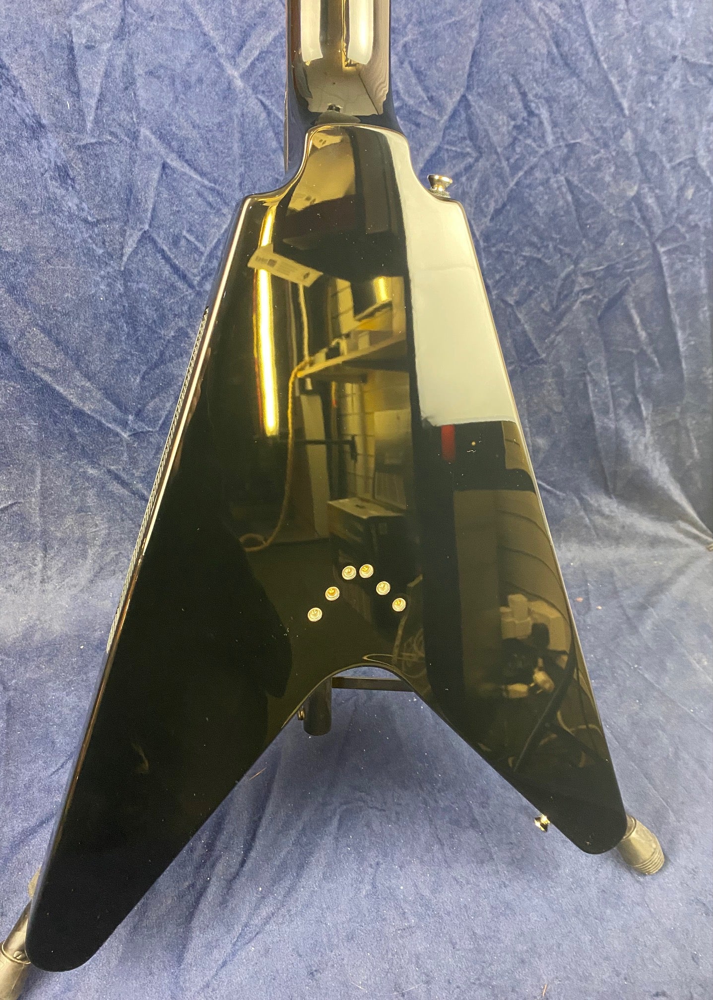 Black V-shaped electric guitar body with a glossy finish, six string ferrules on the back, set against a textured blue background.