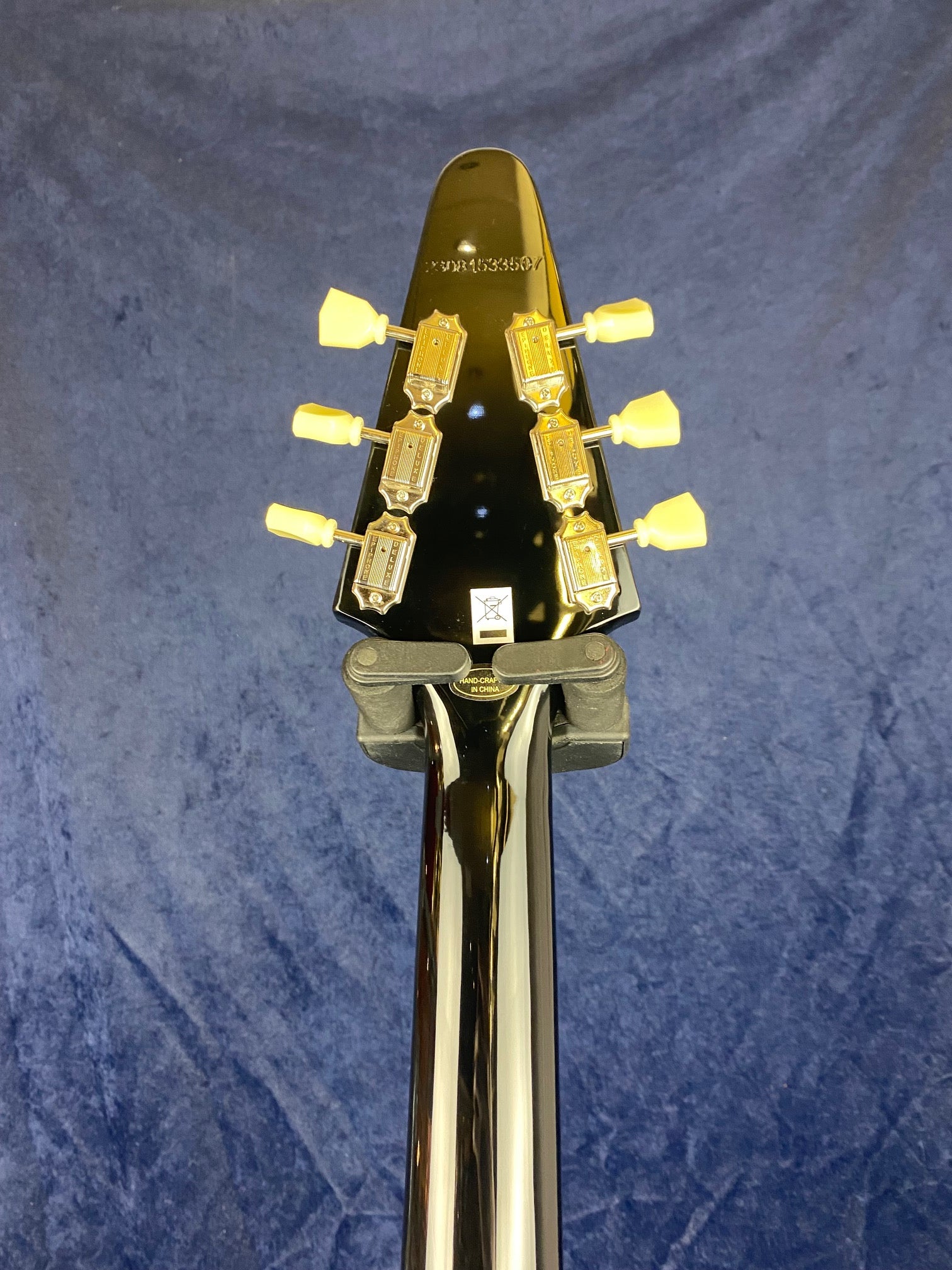 Electric guitar headstock and neck shown from the back, featuring six gold tuning keys with cream buttons against a blue background.