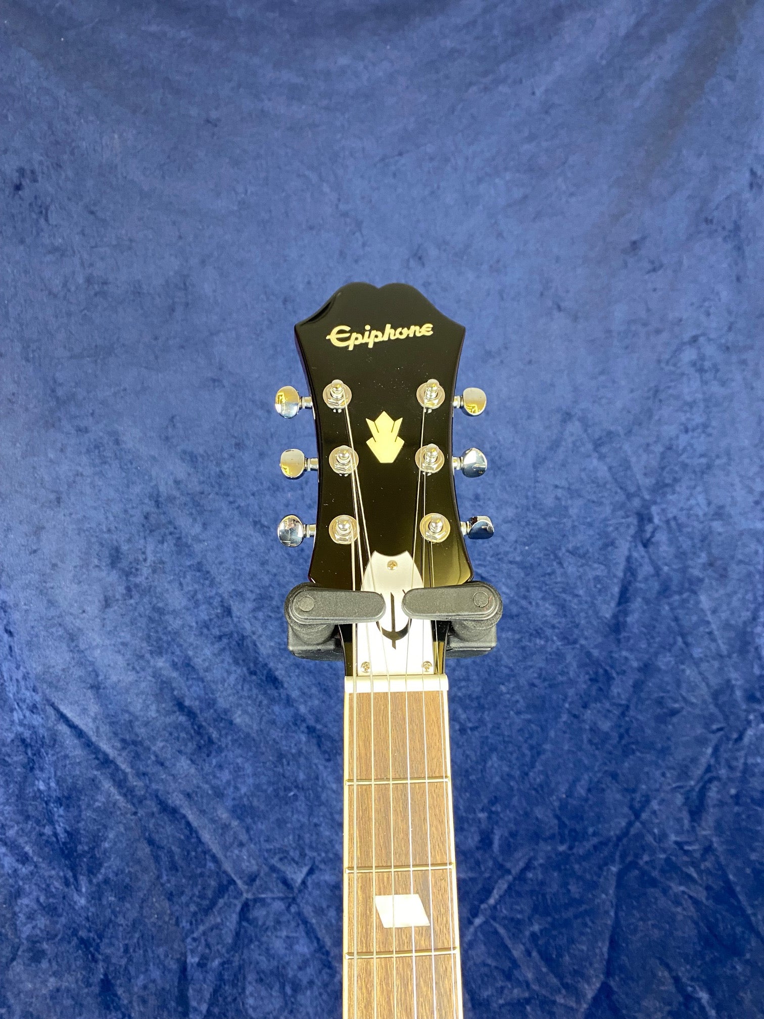 Black Epiphone electric guitar headstock with gold tuners and logo, wooden neck and fretboard, shown upright against a blue textured background.