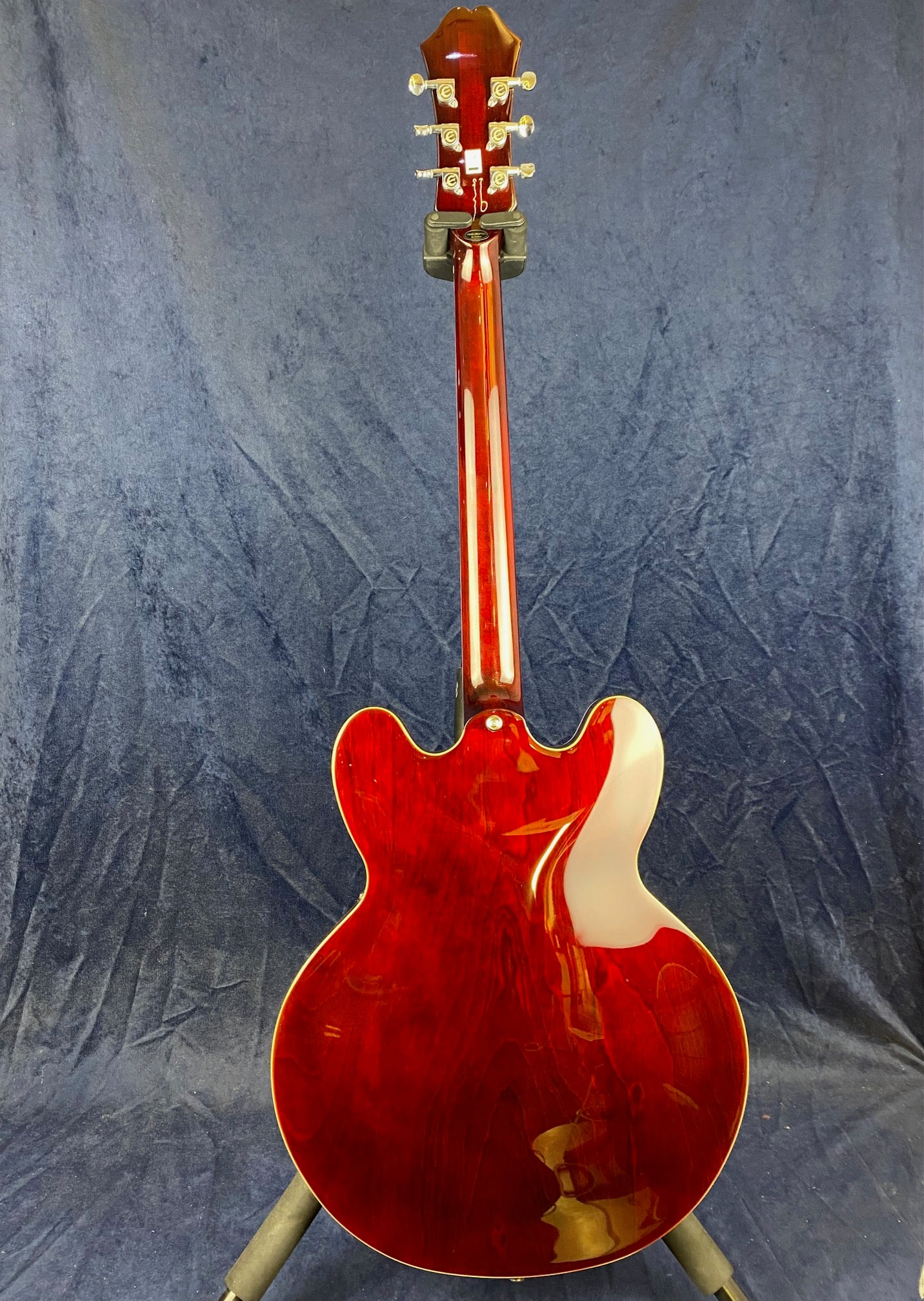 Red semi-hollow electric guitar shown from the back, glossy finish, gold tuning pegs, double cutaway body, propped on a stand against a blue background.