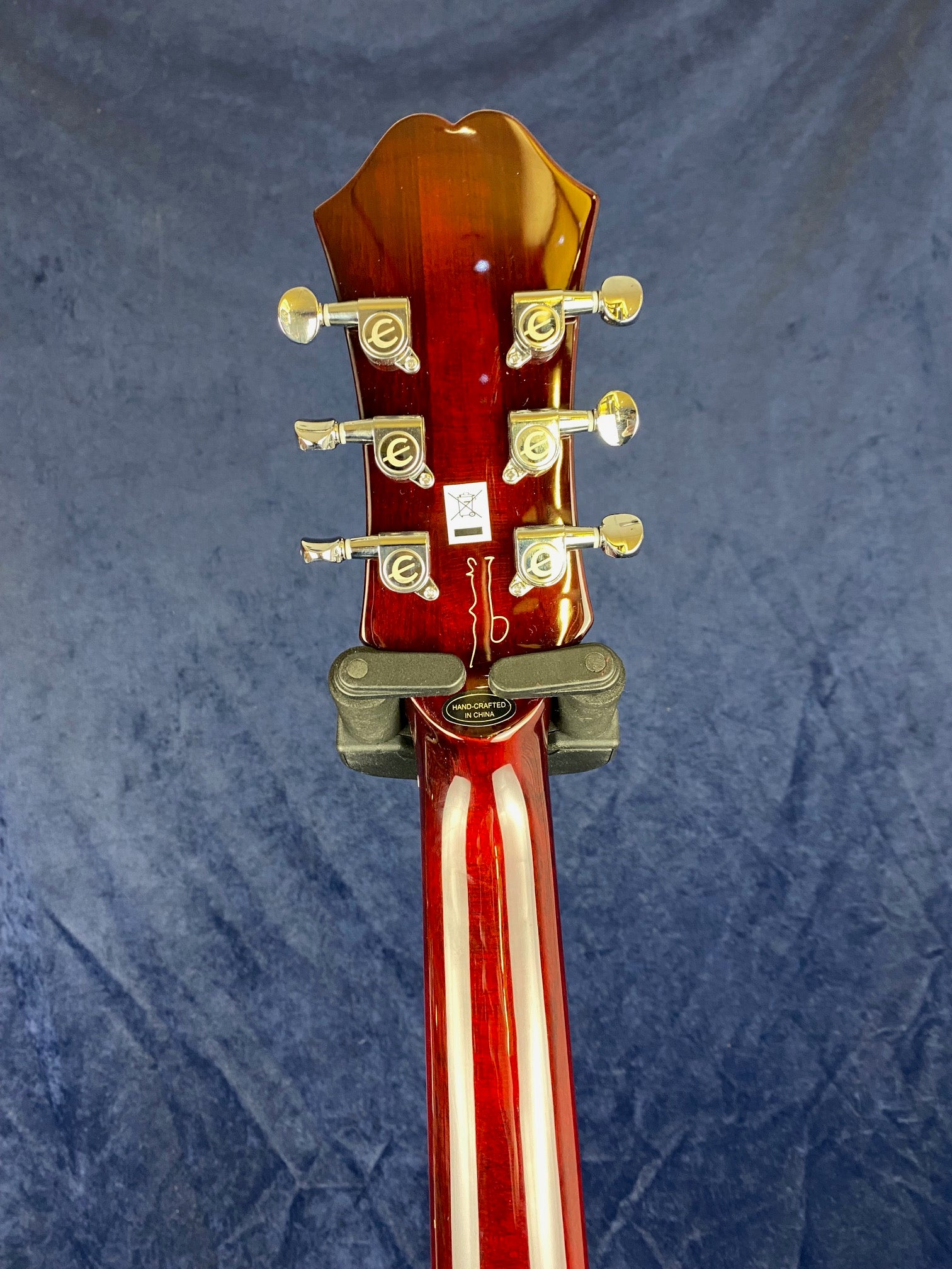 Back of a red electric guitar’s headstock with six gold tuning pegs and a “hand-crafted in China” label on the neck, set against a blue background.