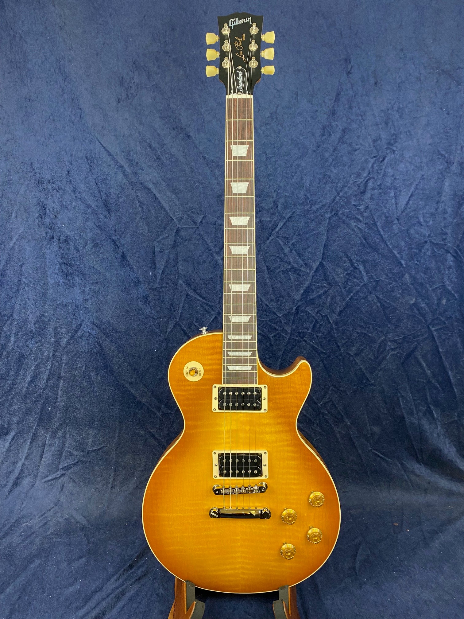 Sunburst Gibson Les Paul electric guitar with two humbucker pickups, four control knobs, and a single cutaway body shape against a dark blue background.