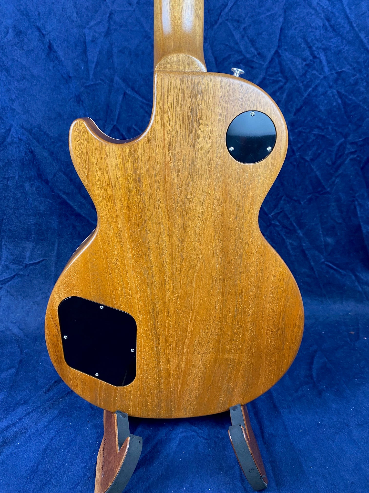 The back of a brown wood electric guitar with two black plastic access plates, shown on a stand against a dark blue fabric background.