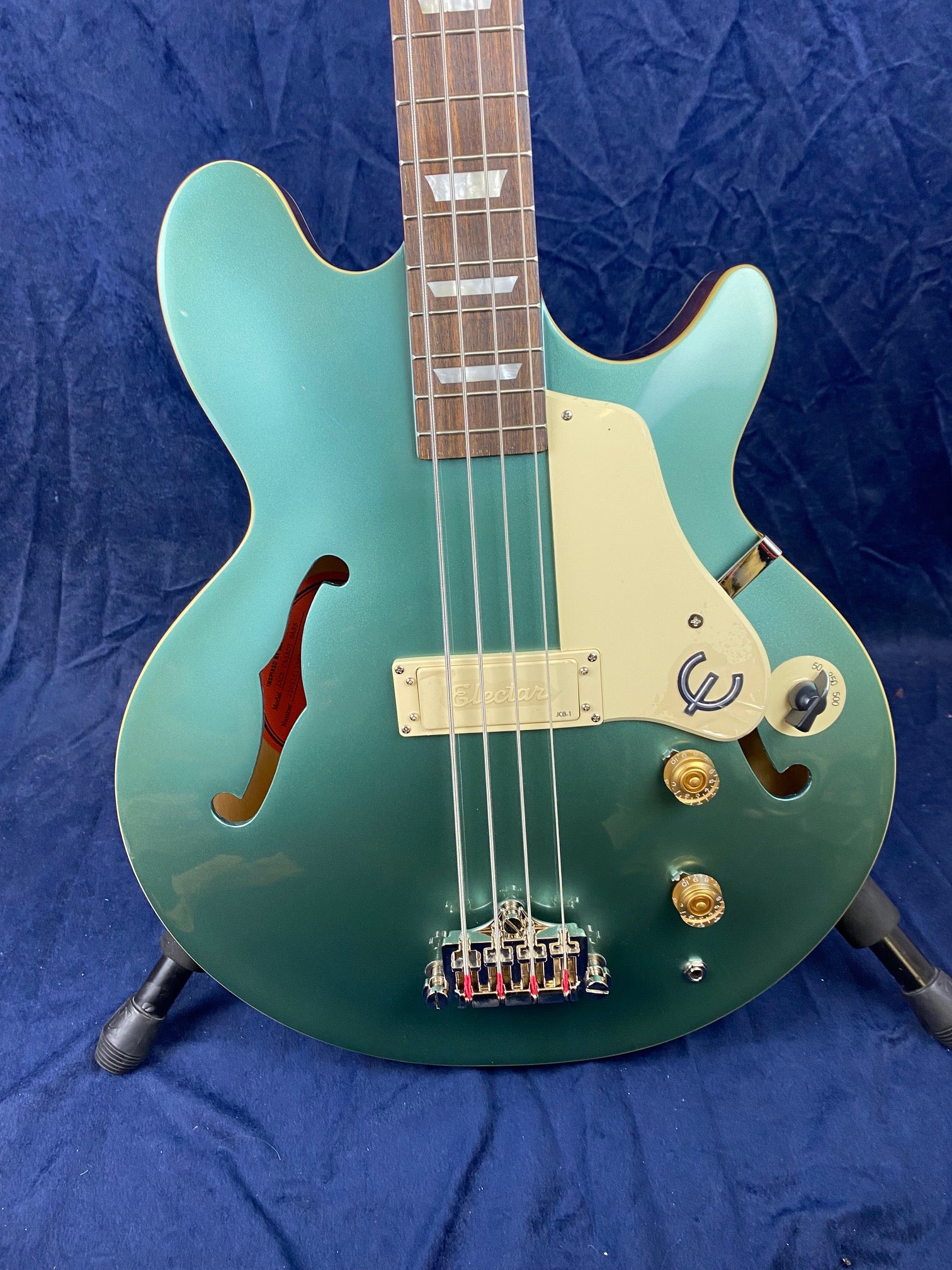 Seafoam green semi-hollow bass guitar with f-holes, gold knobs, cream pickguard, and four strings, resting on a stand against a blue background.