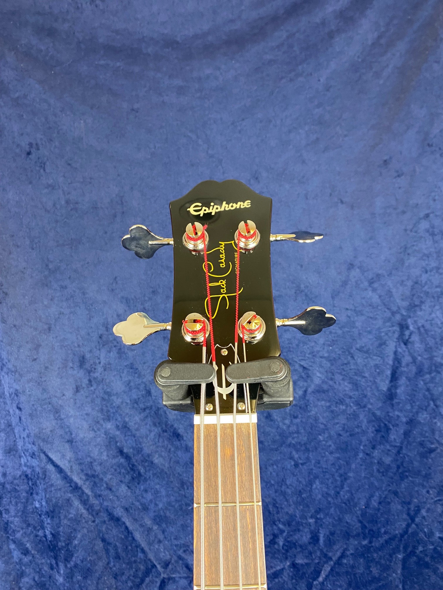 Epiphone bass guitar headstock with four tuning pegs, red string ends, and a signature in yellow and red on a blue background.