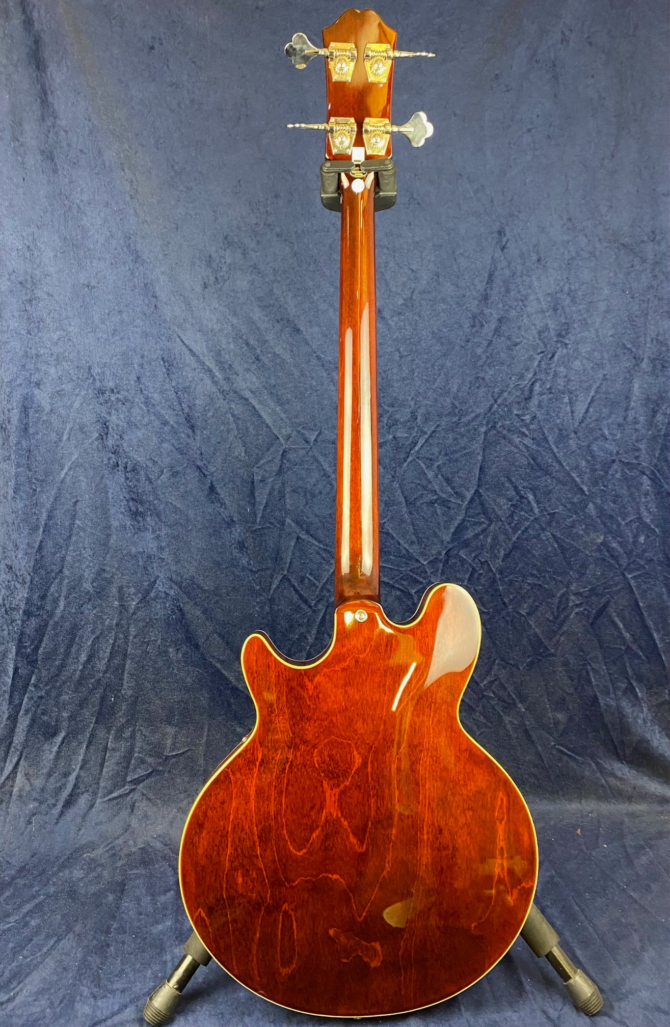 Glossy brown bass guitar with a double-cutaway body shown from the back, silver tuners, and a wood grain finish, resting on a stand.