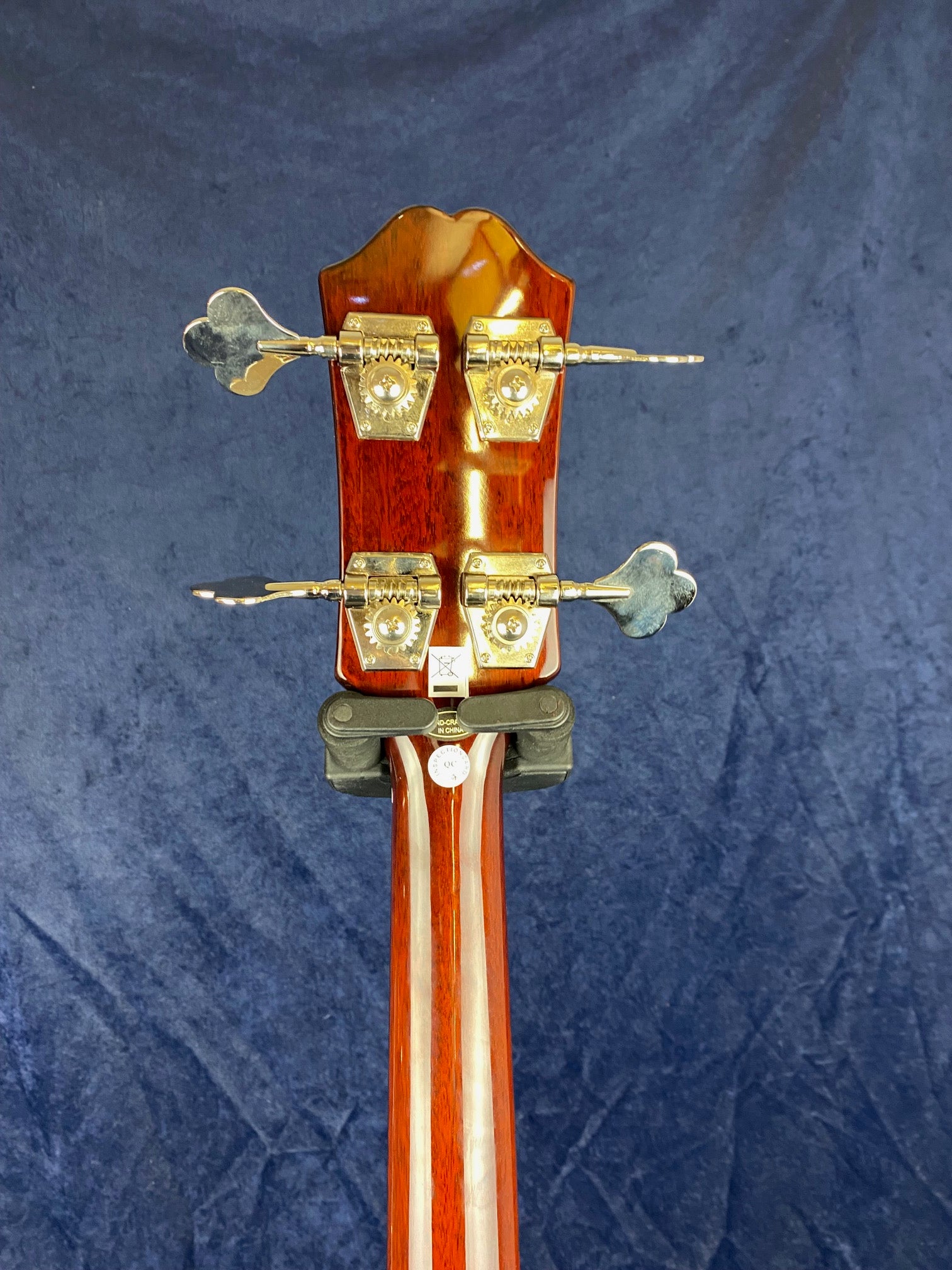Rear view of a bass guitar headstock showing four gold tuning pegs on a glossy, multi-piece wooden neck against a dark blue background.