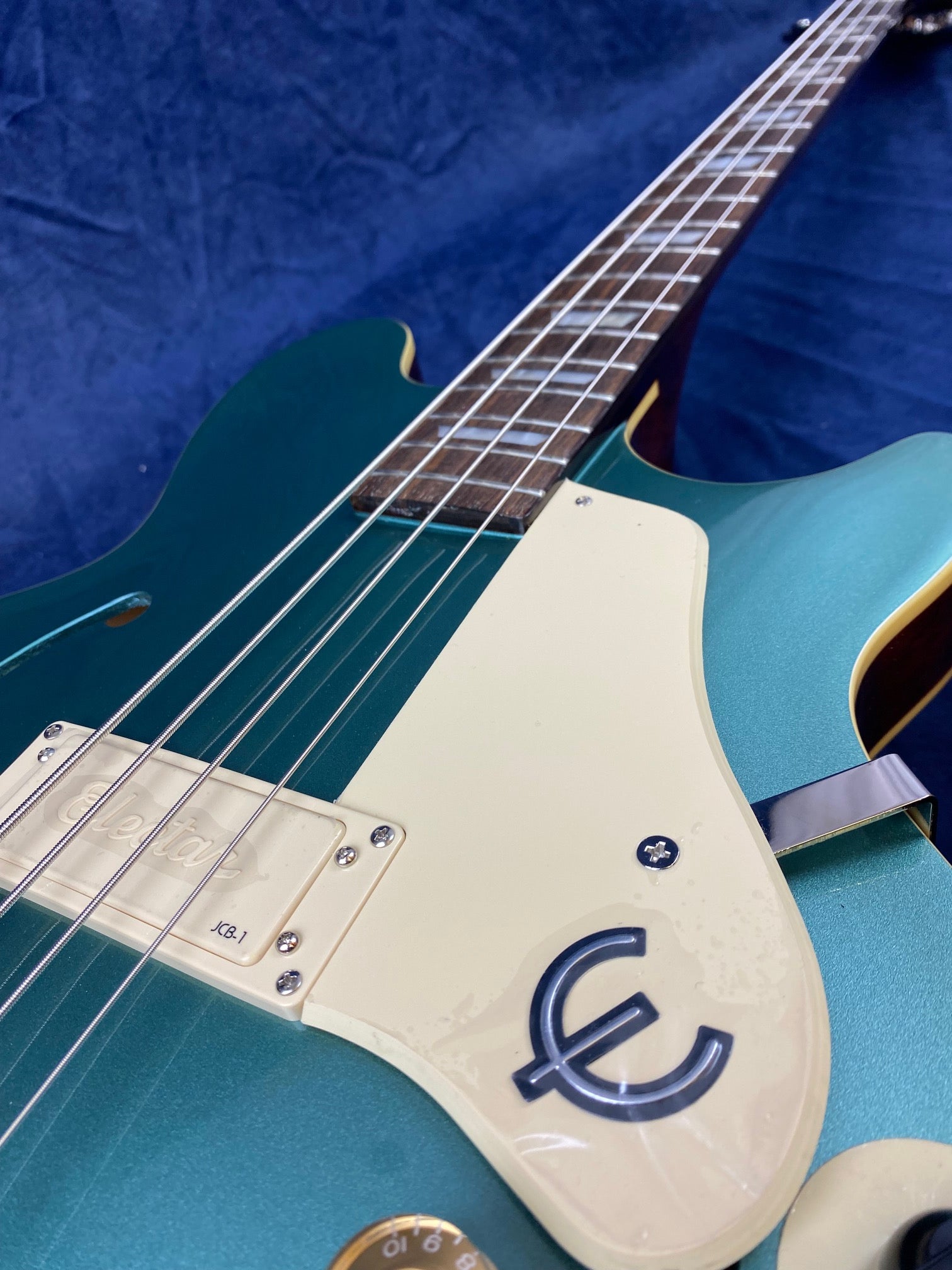 A teal Epiphone bass guitar with cream pickguard, chrome pickup cover, and visible strings, photographed close-up against a dark blue background.