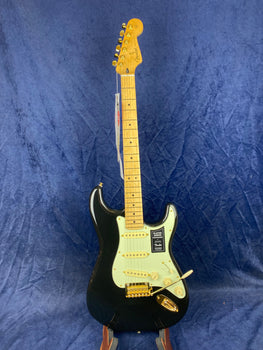 Black Fender electric guitar with a light-colored pickguard, gold hardware, and maple neck, standing upright against a dark blue background.