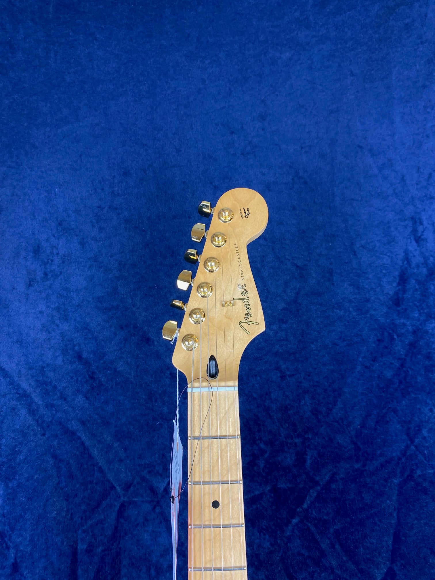 Light-colored Fender electric guitar neck and headstock with six gold tuning pegs, set against a textured blue background.