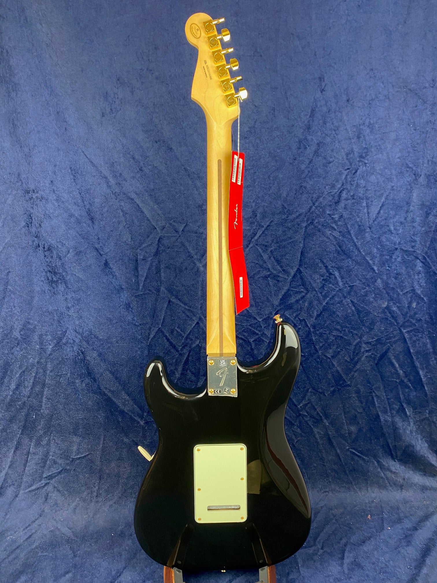 Black electric guitar seen from the back, maple neck, gold hardware, white backplate, and red Fender tags hanging from a tuning peg.