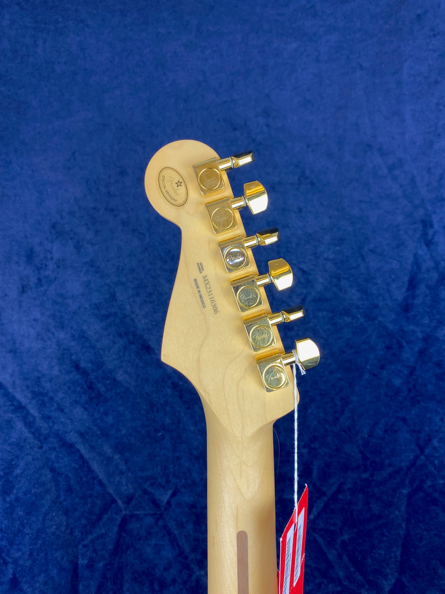 The back of an electric guitar headstock with six gold tuning pegs and attached red tags, against a solid dark blue background.