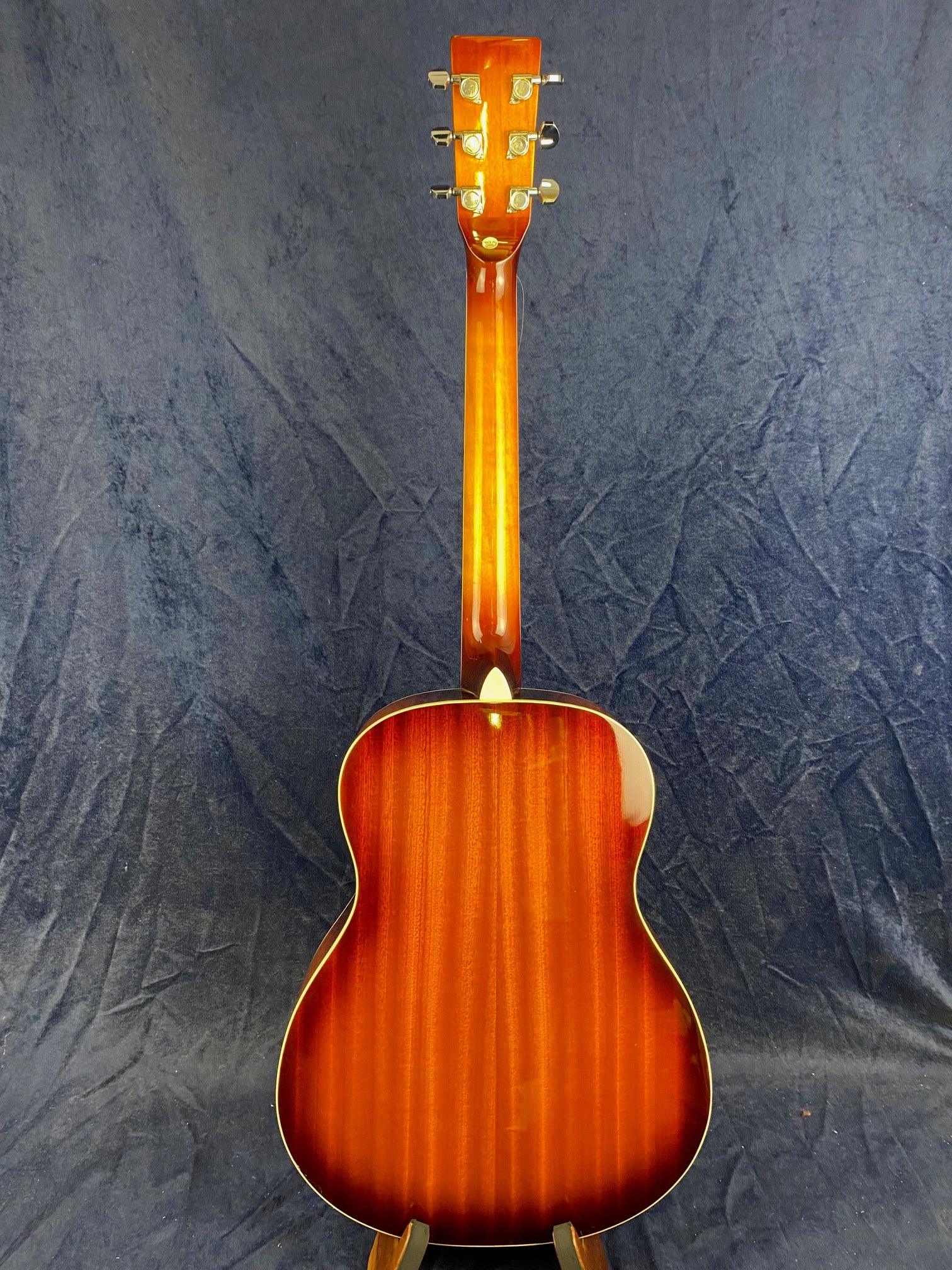 A resonator guitar shown from the back, featuring a glossy reddish-brown wood finish with gold tuning pegs, set against a dark textured background.