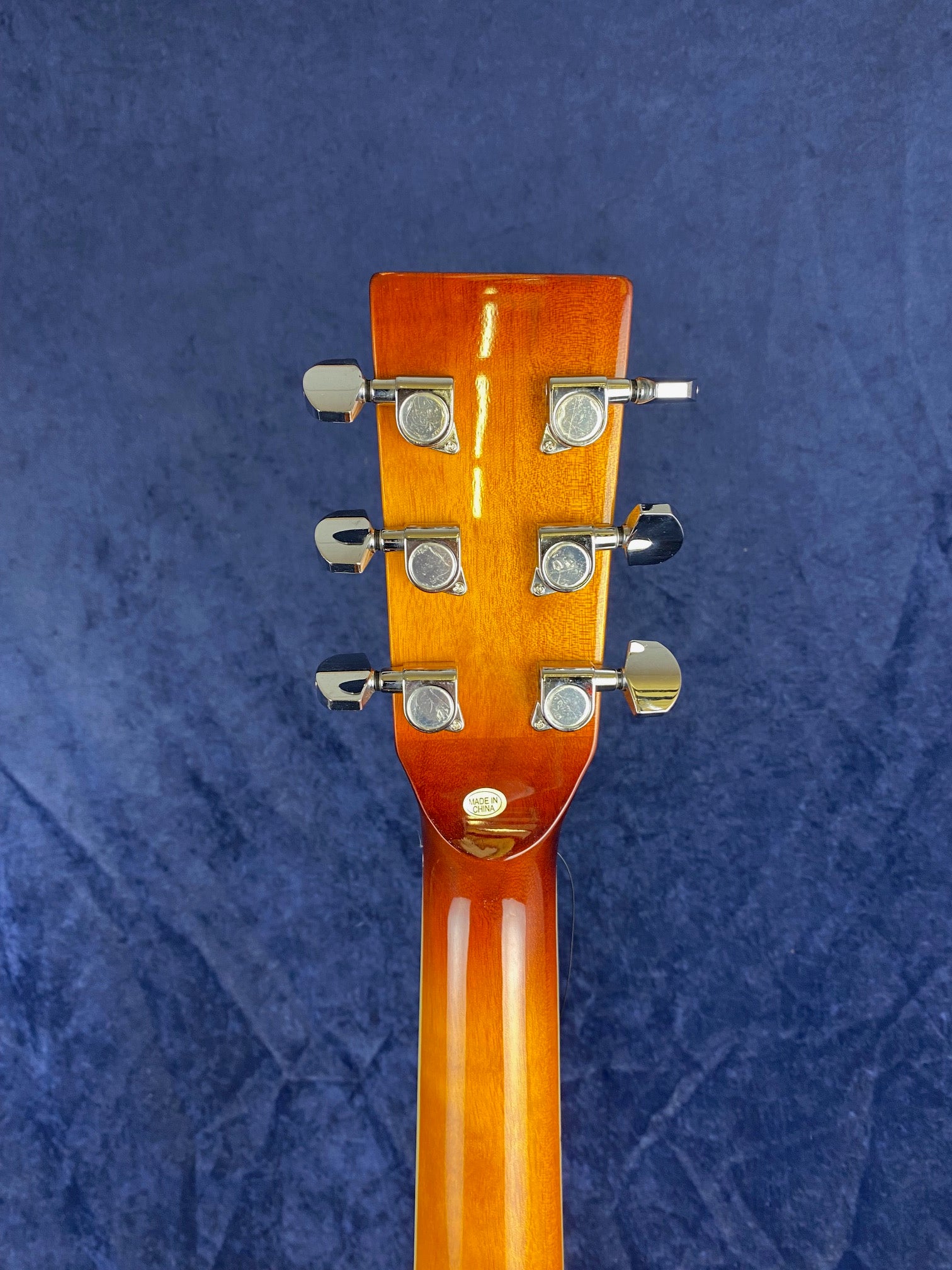 Back of a resonator guitar headstock showing six chrome tuning pegs, a glossy wood finish, and a “Made in China” sticker at the base.