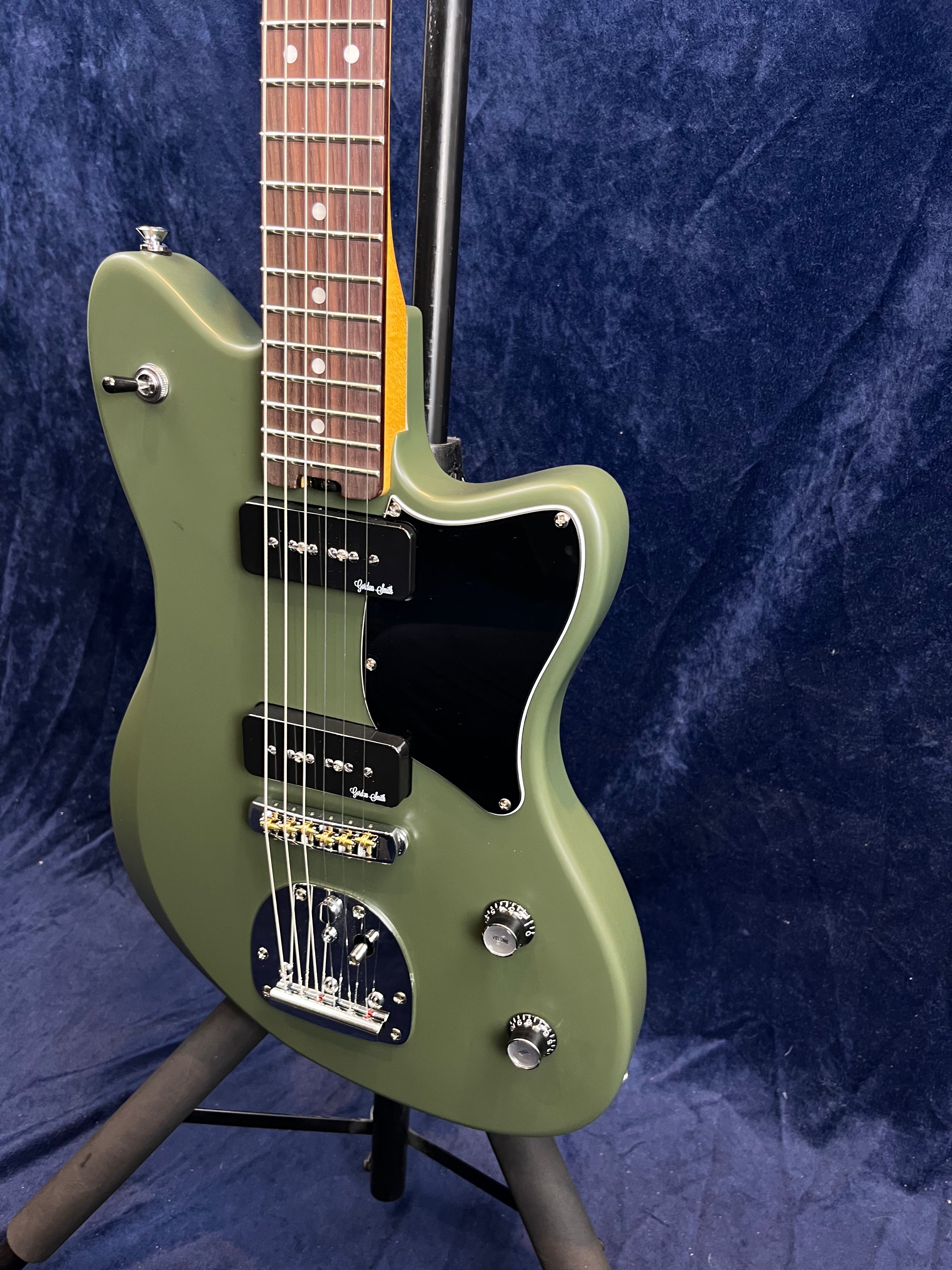 Matte olive green electric guitar with two black pickups, black pickguard, chrome bridge, and two control knobs, shown upright on a stand.
