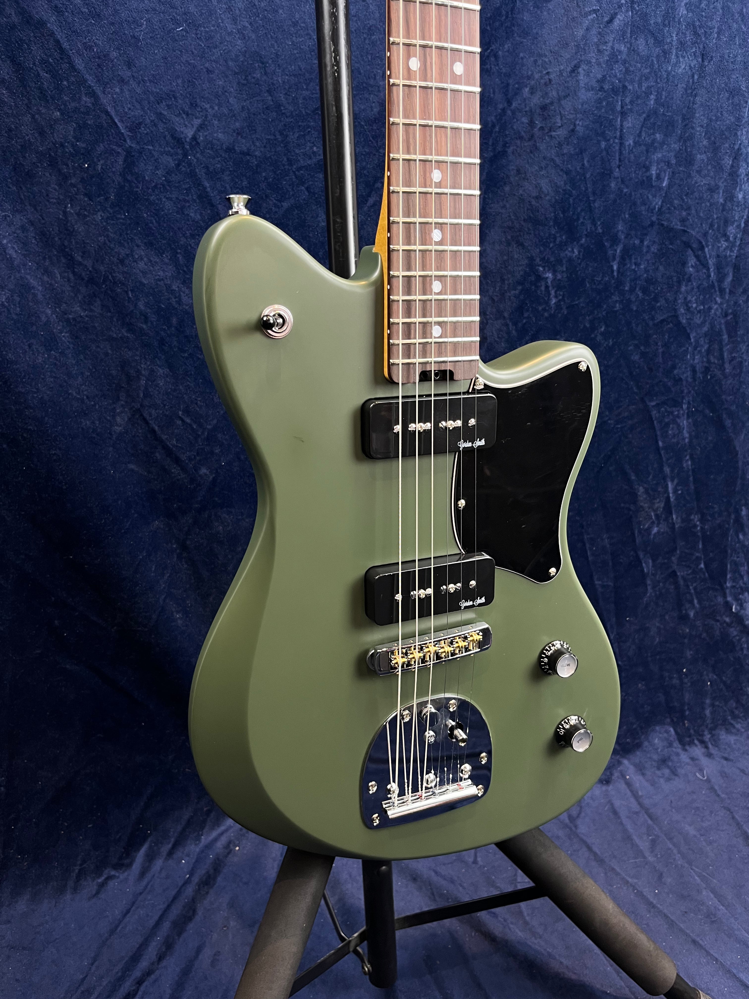 Matte olive green electric guitar with two black pickups, black pickguard, chrome hardware, and two control knobs, resting on a stand.