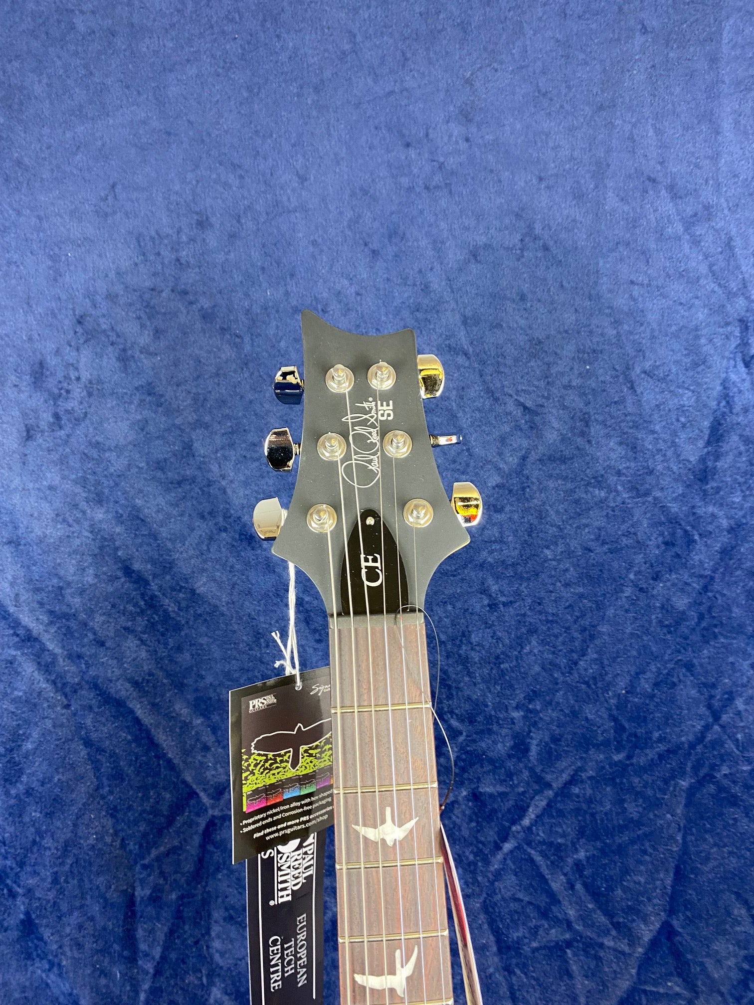 Headstock and upper neck of a PRS electric guitar with gold tuning pegs and bird inlays, set against a blue background, tags attached to the headstock.