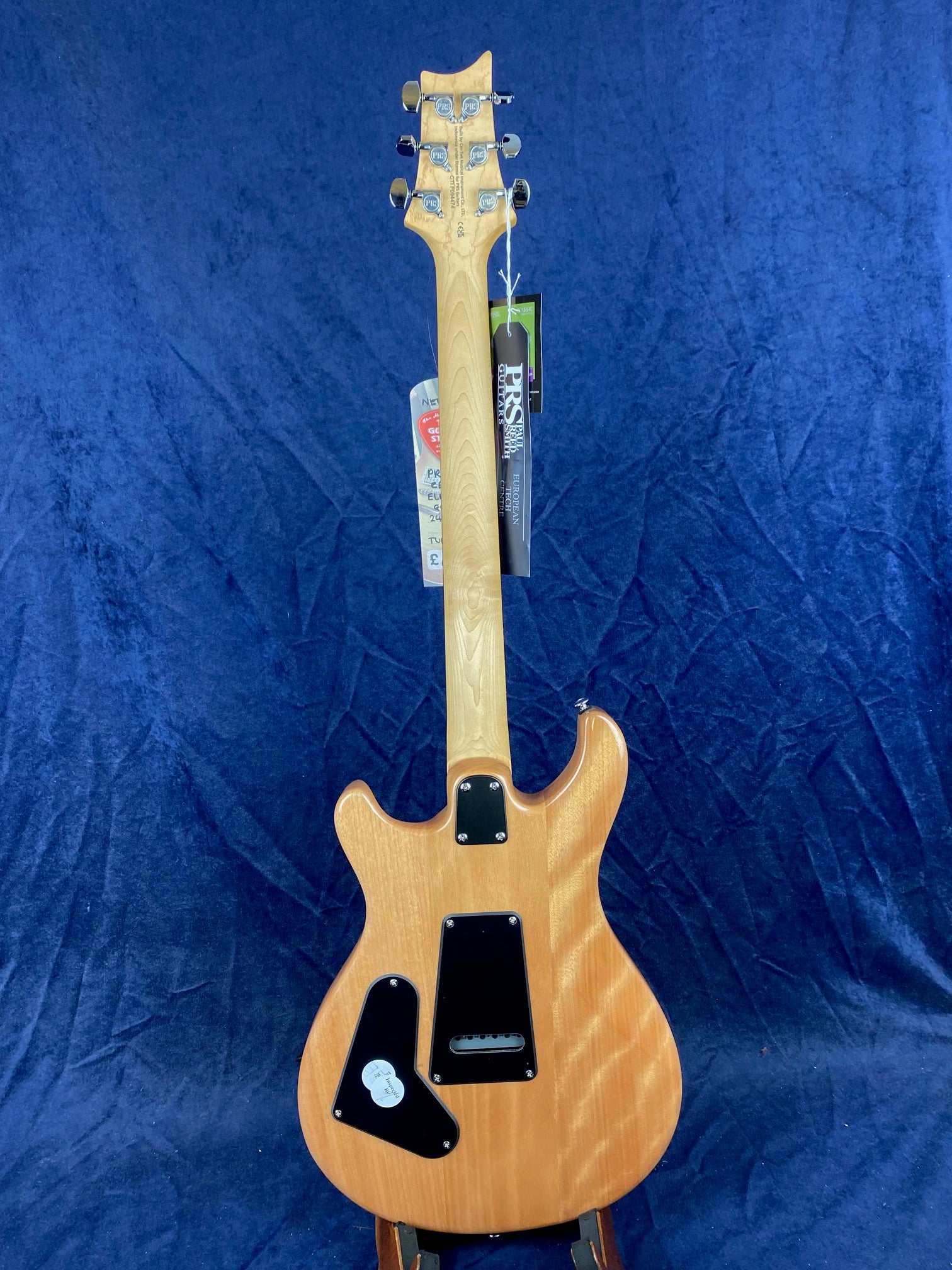 Natural wood finish electric guitar shown from the back with three black plastic cover plates and silver tuning pegs, tags hanging from the headstock.