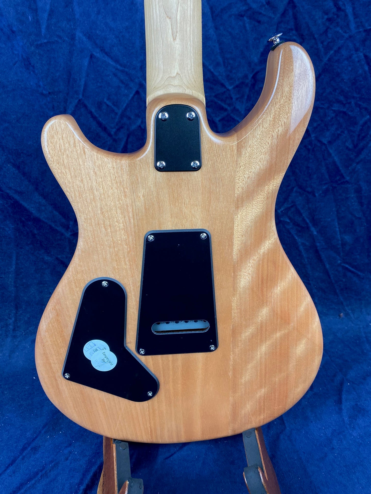 Natural wood electric guitar body with three black plastic cover plates and screws on the back, displayed vertically against a dark blue background.