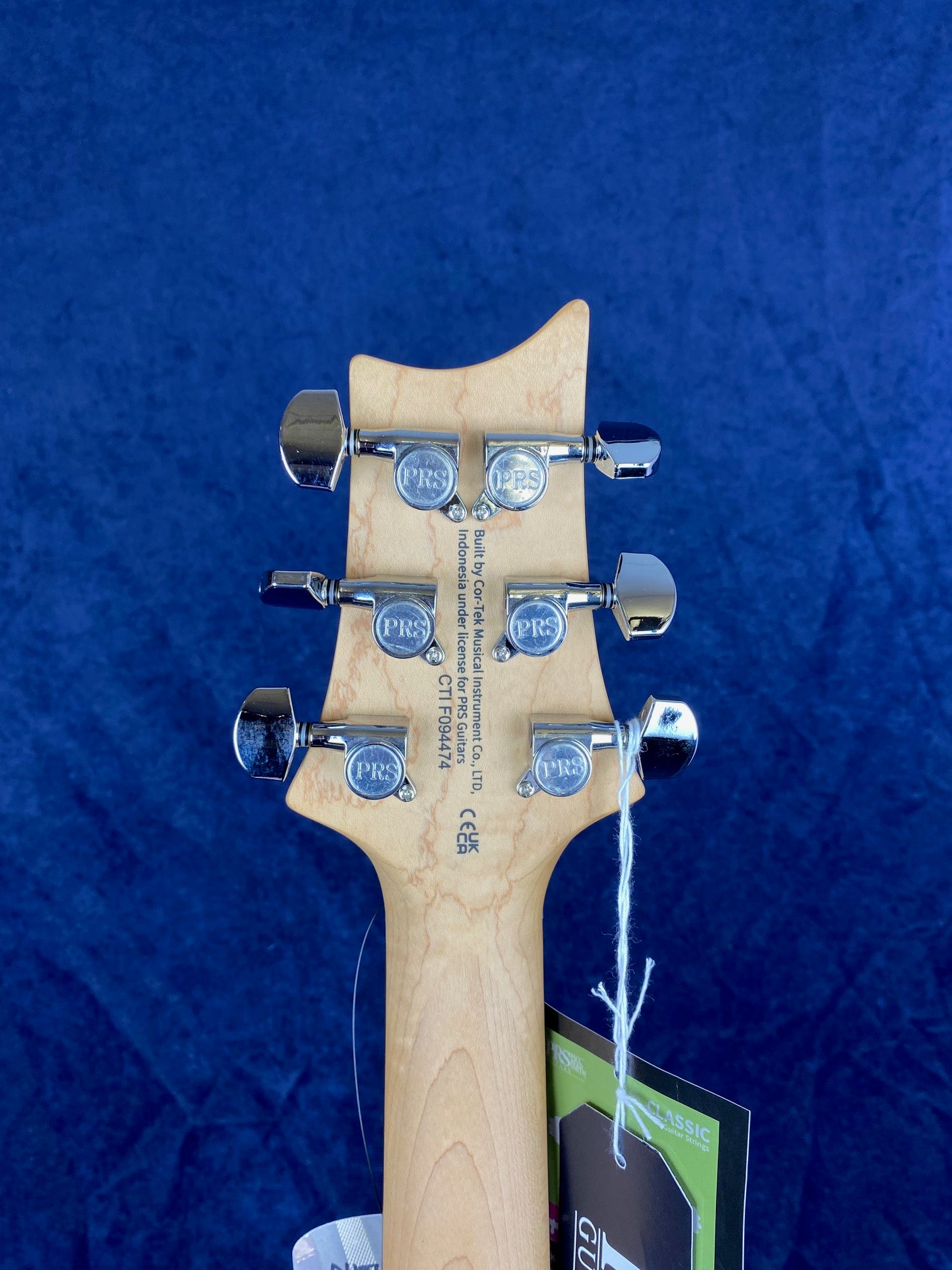 Light wood electric guitar headstock with six chrome PRS tuners, tags attached, and product info stamped on the back against a blue background.